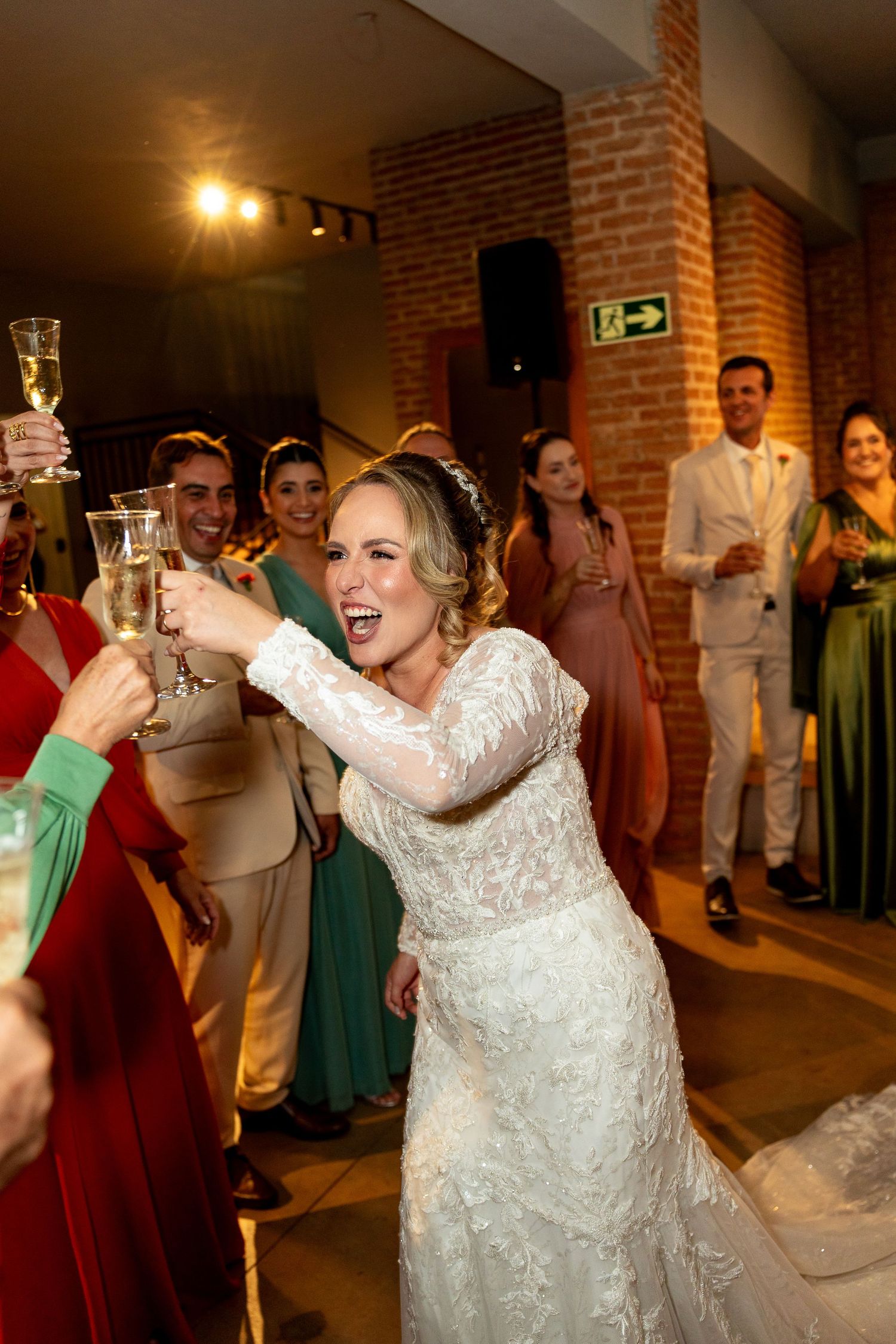 Bride in lace gown toasting champagne with guests at wedding reception, bridesmaids in green dresses cheering behind her.