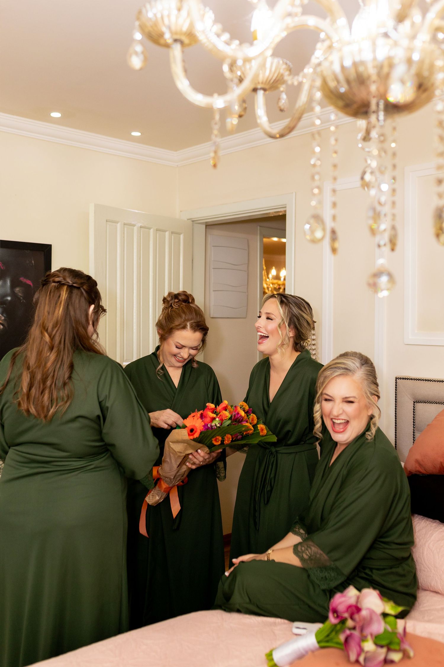 Four bridesmaids in green robes laughing and admiring a vibrant orange bouquet in an elegant room with a crystal chandelier.