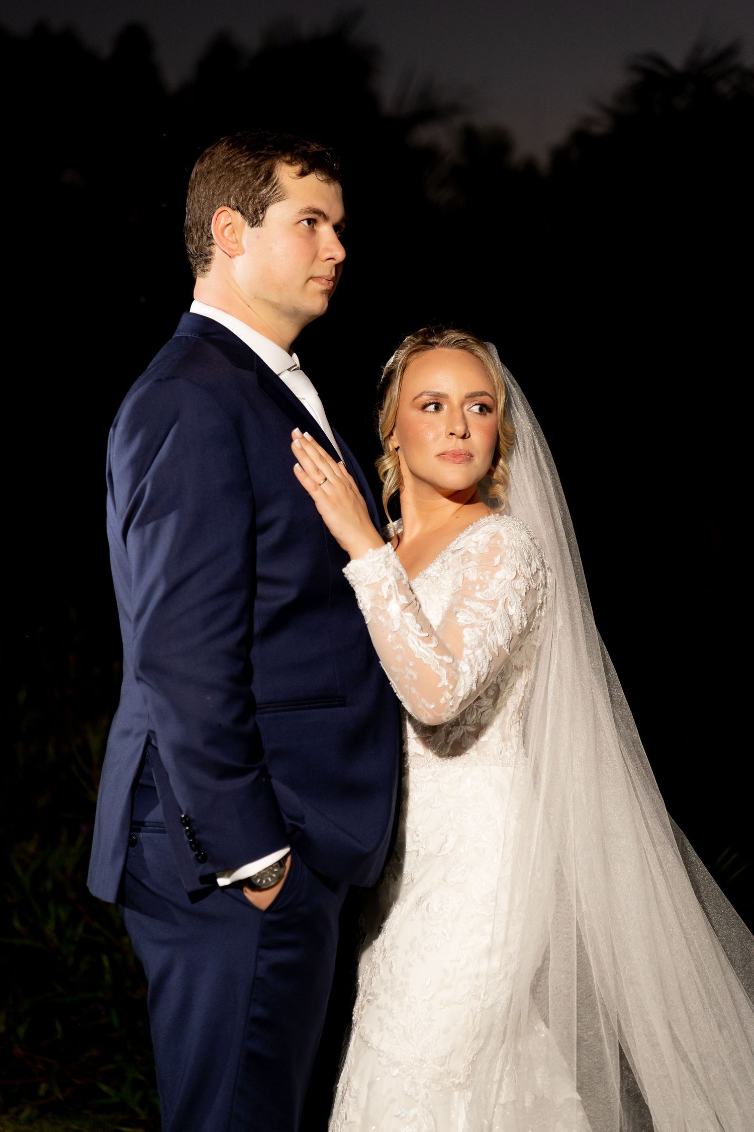 Bride in lace gown and veil stands beside groom in navy suit during elegant outdoor nighttime wedding portrait.
