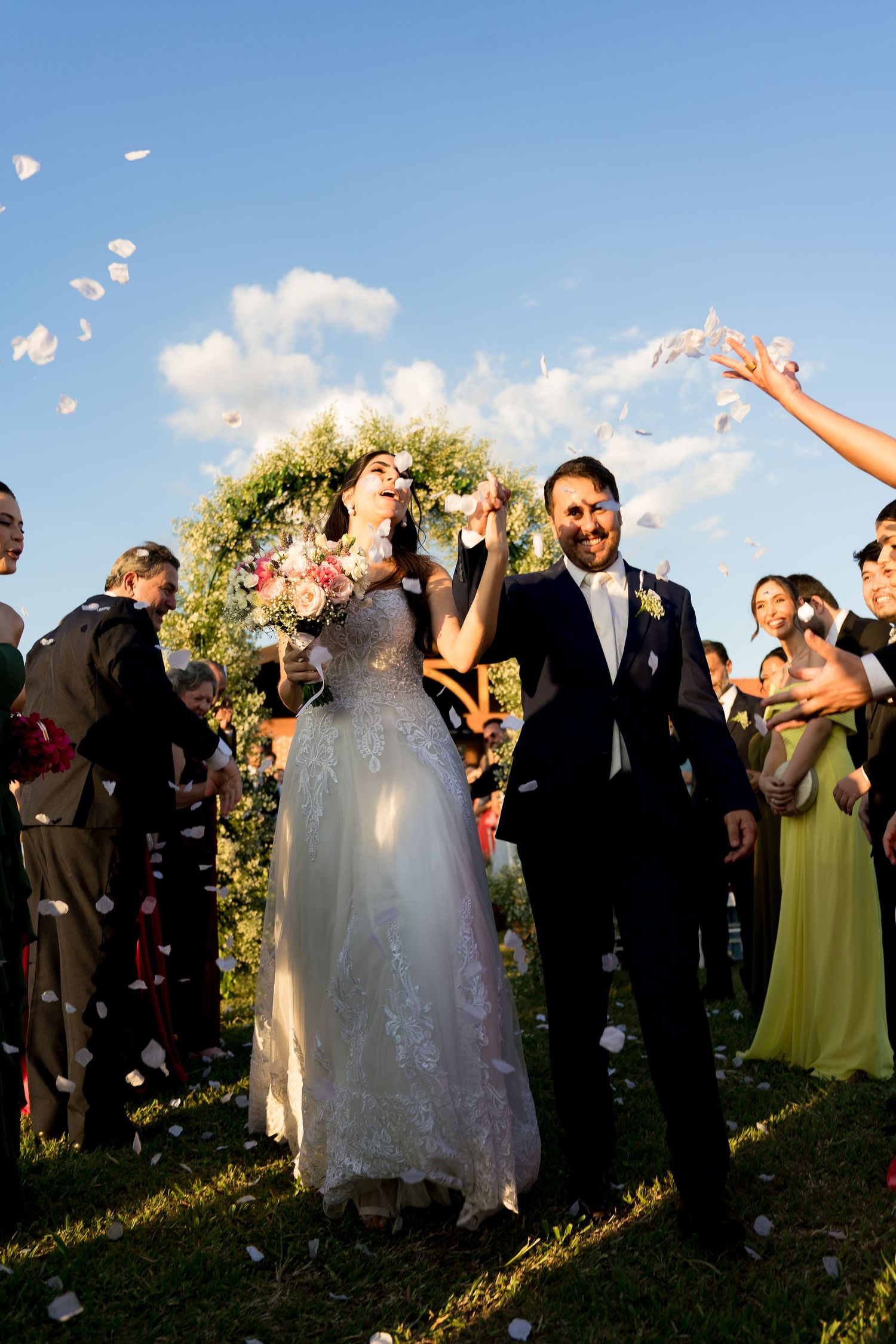 Bride and groom walk through flower petal confetti after outdoor wedding ceremony under blue sky.