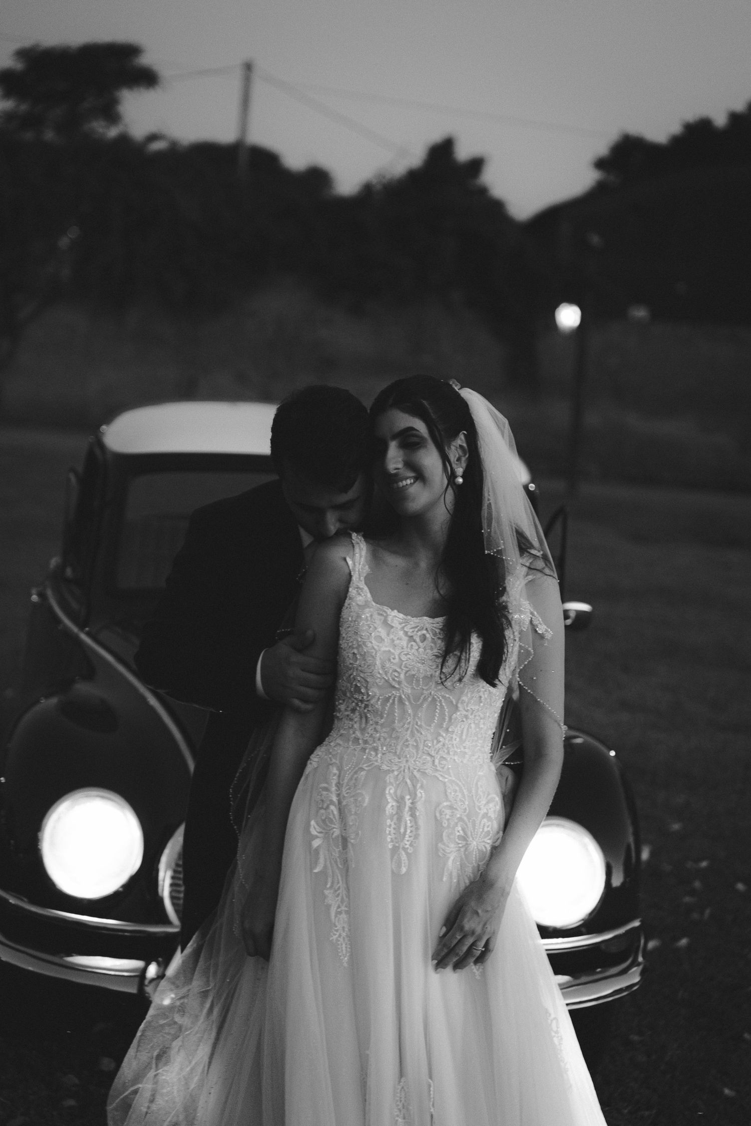 Black and white photo of a smiling bride in a lace gown and veil with groom beside a vintage car at dusk.