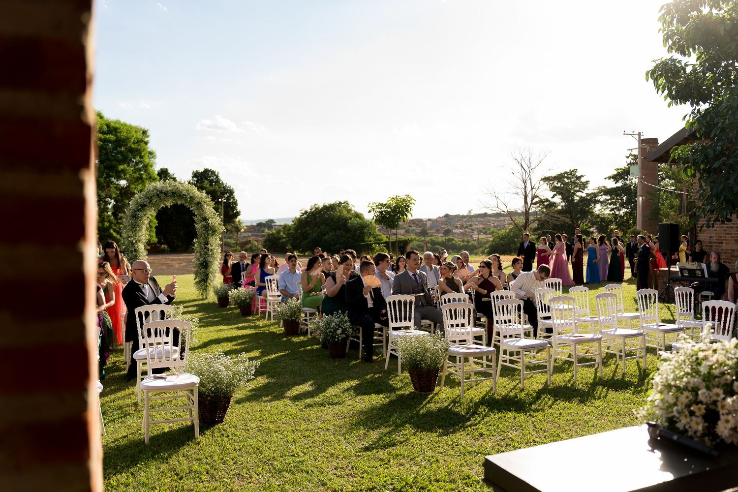 Outdoor wedding ceremony on green lawn with white chairs and guests gathered under open sky at a rustic venue.