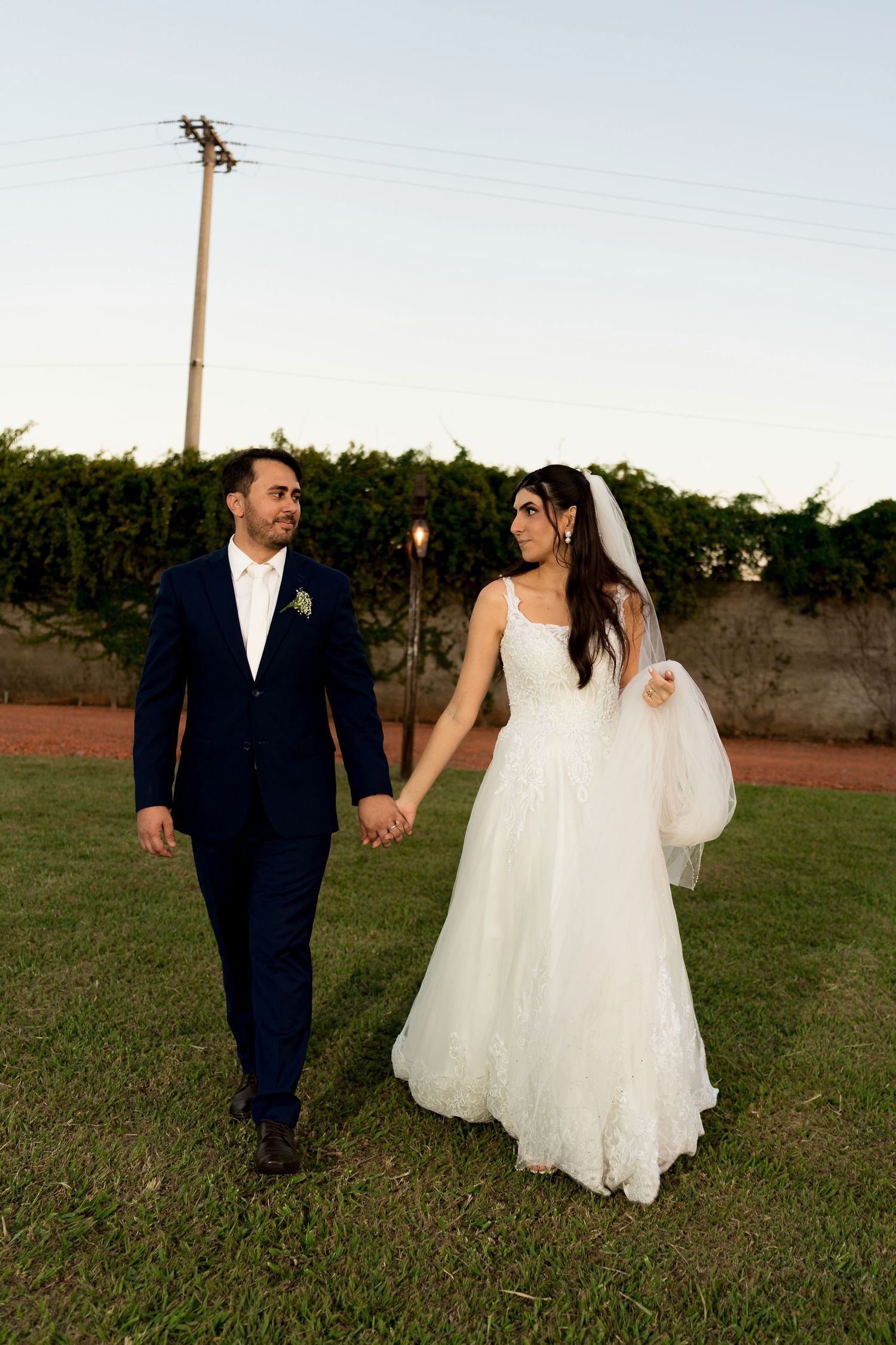 Bride in white gown and groom in navy suit hold hands walking on green lawn at outdoor wedding.