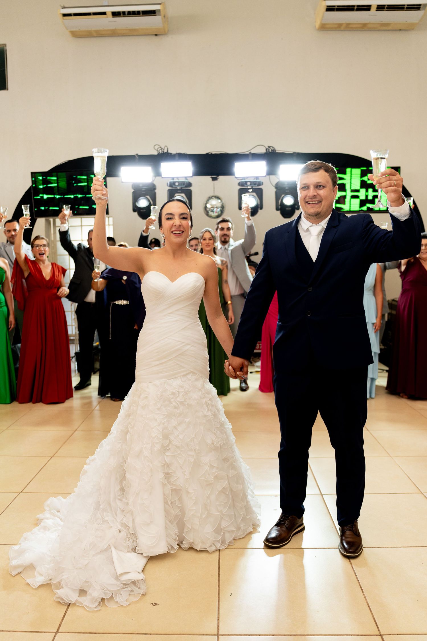 Bride in white gown and groom in navy suit hold hands and raise glasses on wedding dance floor with guests behind them.
