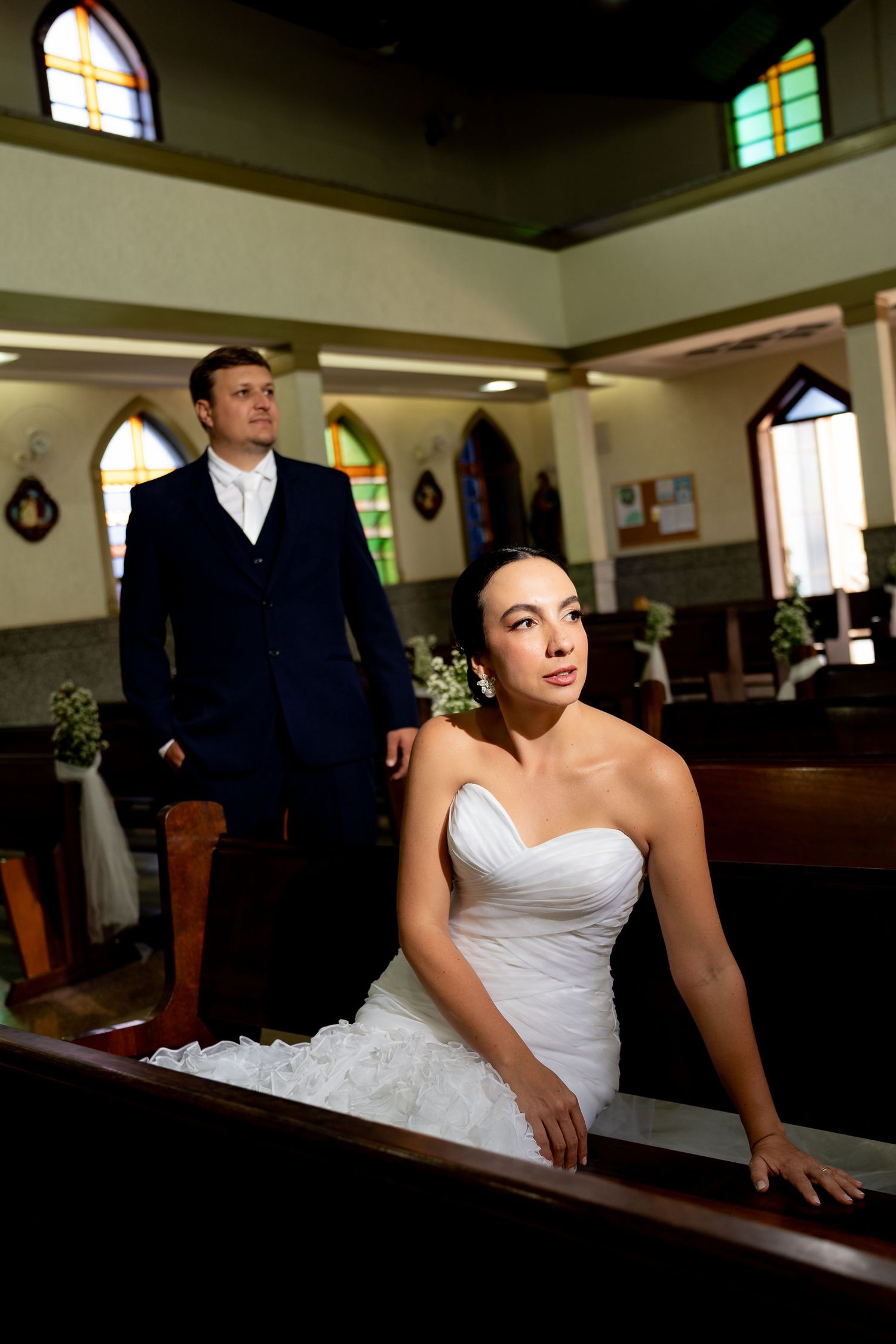 Bride in white strapless gown sits in church pew while groom in navy suit stands behind her during wedding ceremony.