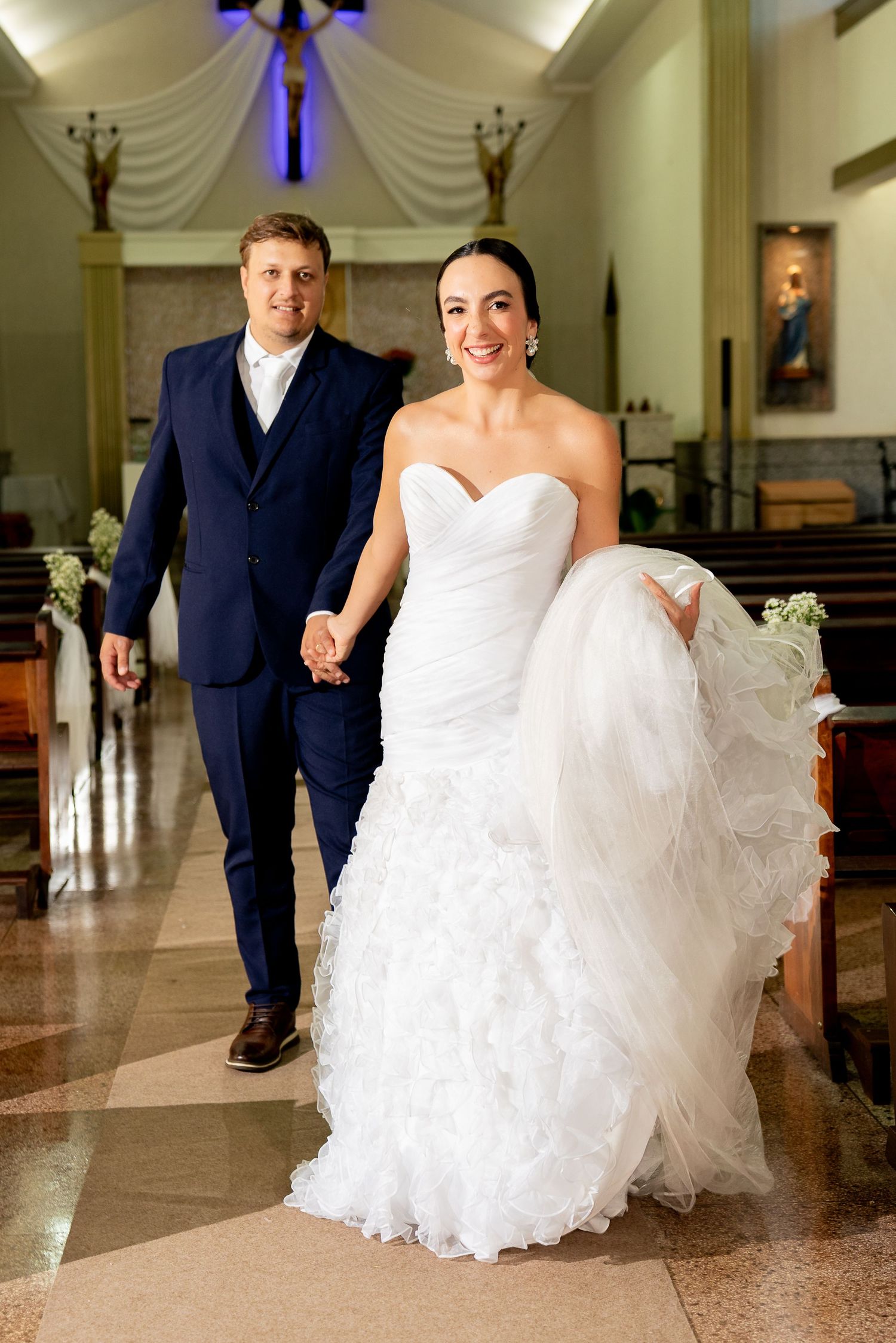 Bride in white mermaid gown and groom in navy suit walk hand in hand down church aisle after wedding ceremony.