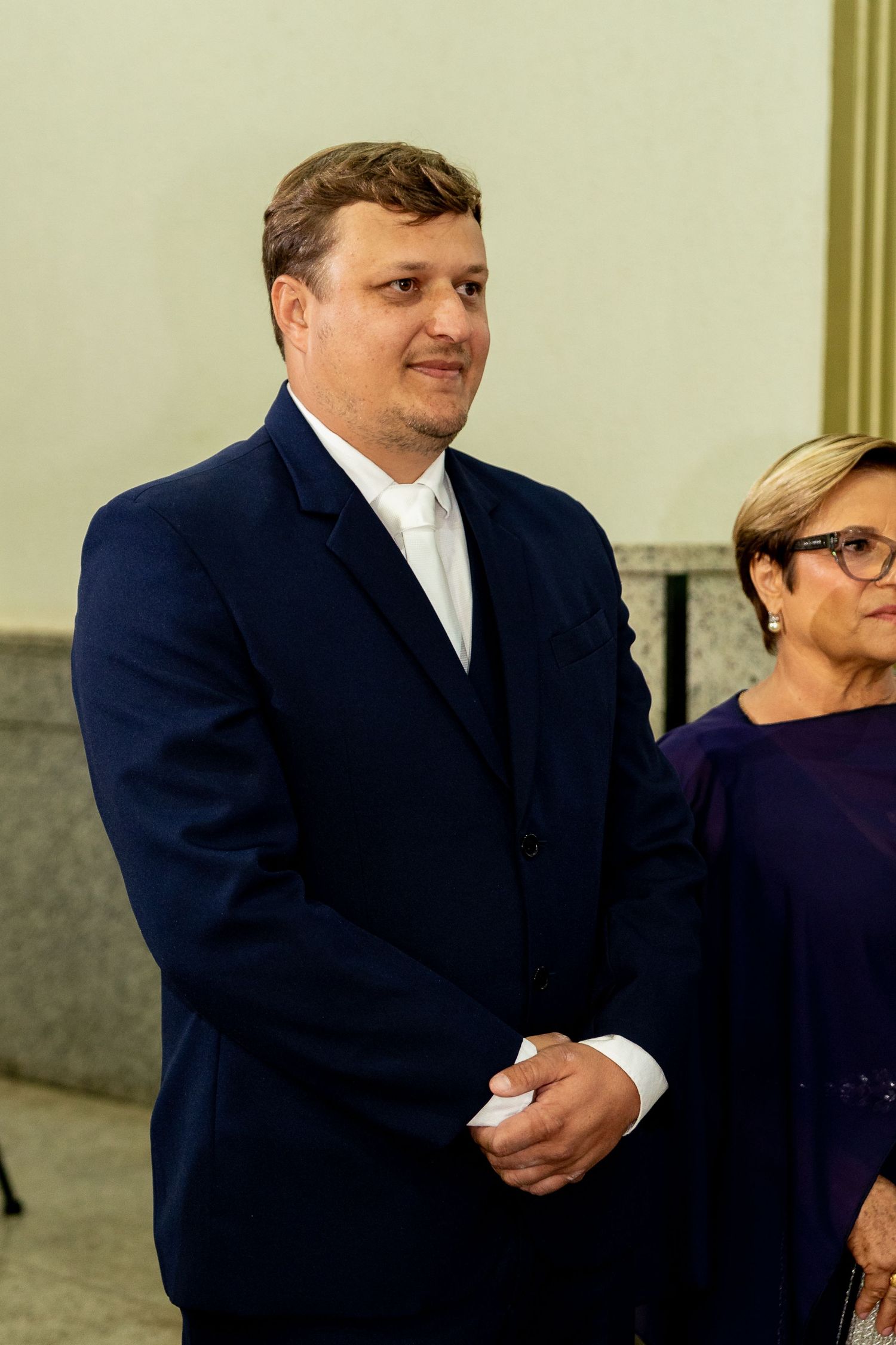 Man in navy blue suit smiling at formal event, standing beside woman in purple dress.