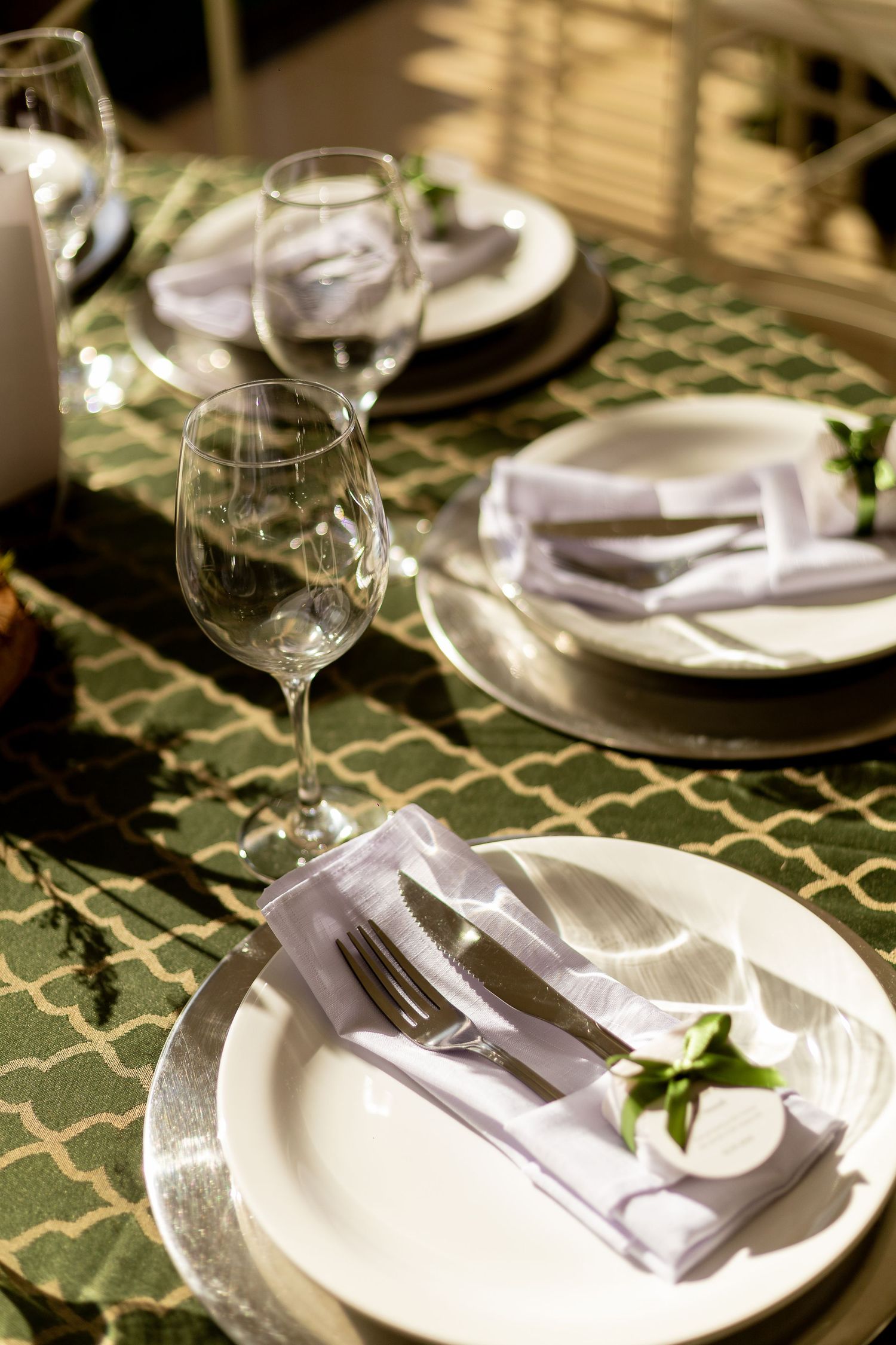 Elegant table setting with white plates, silver chargers, folded napkins, and crystal wine glasses on a green patterned tablecloth.