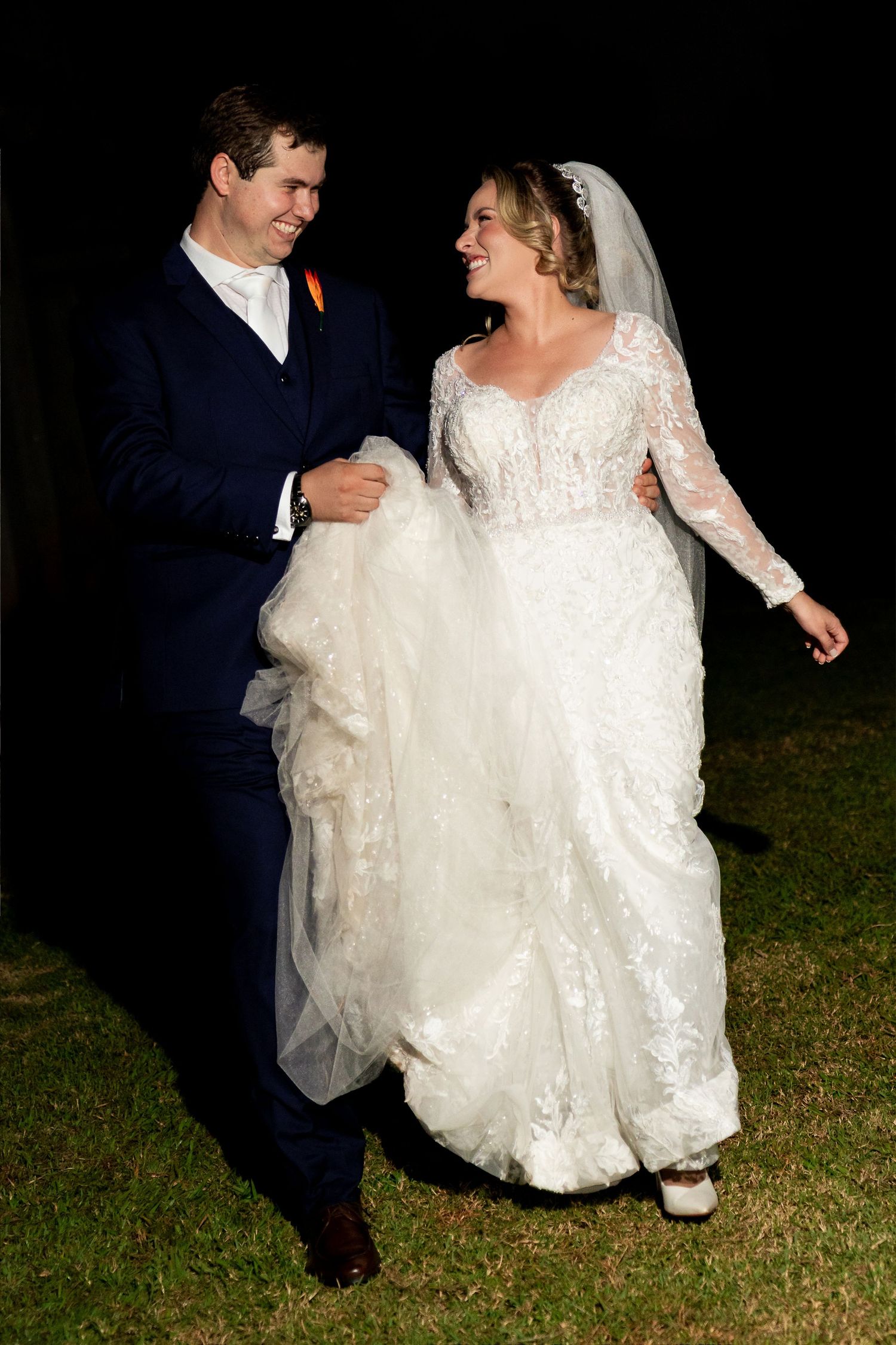 Happy bride and groom walking together outdoors at night, bride in lace wedding gown with veil, groom in navy suit.