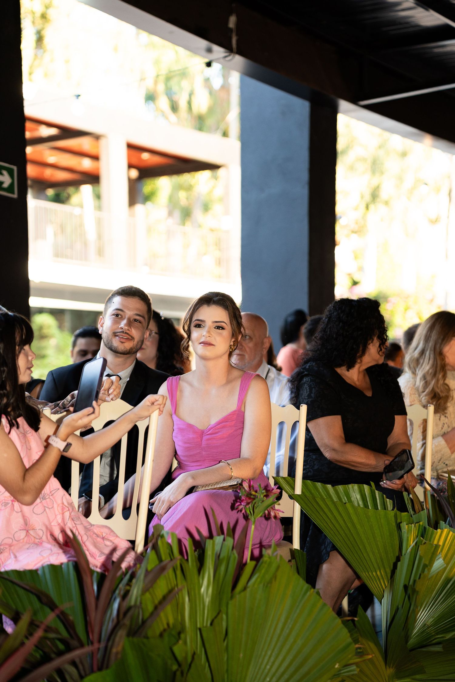 Guests seated at an outdoor wedding ceremony, woman in pink dress smiling, man in suit beside her, tropical greenery in foreground.