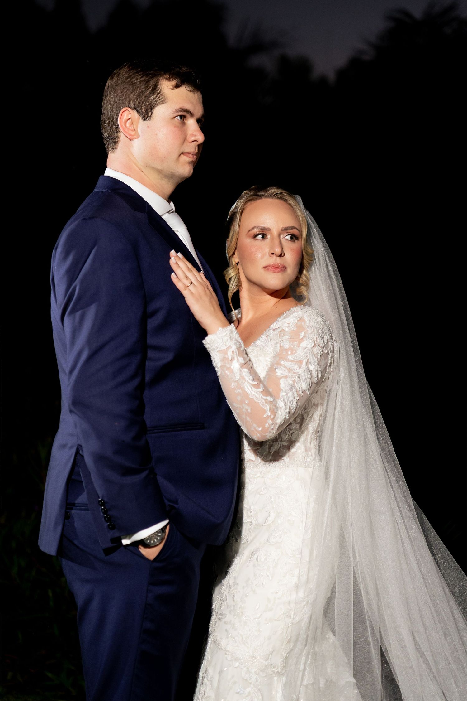 Bride in lace gown and veil stands beside groom in navy suit during elegant nighttime wedding portrait.