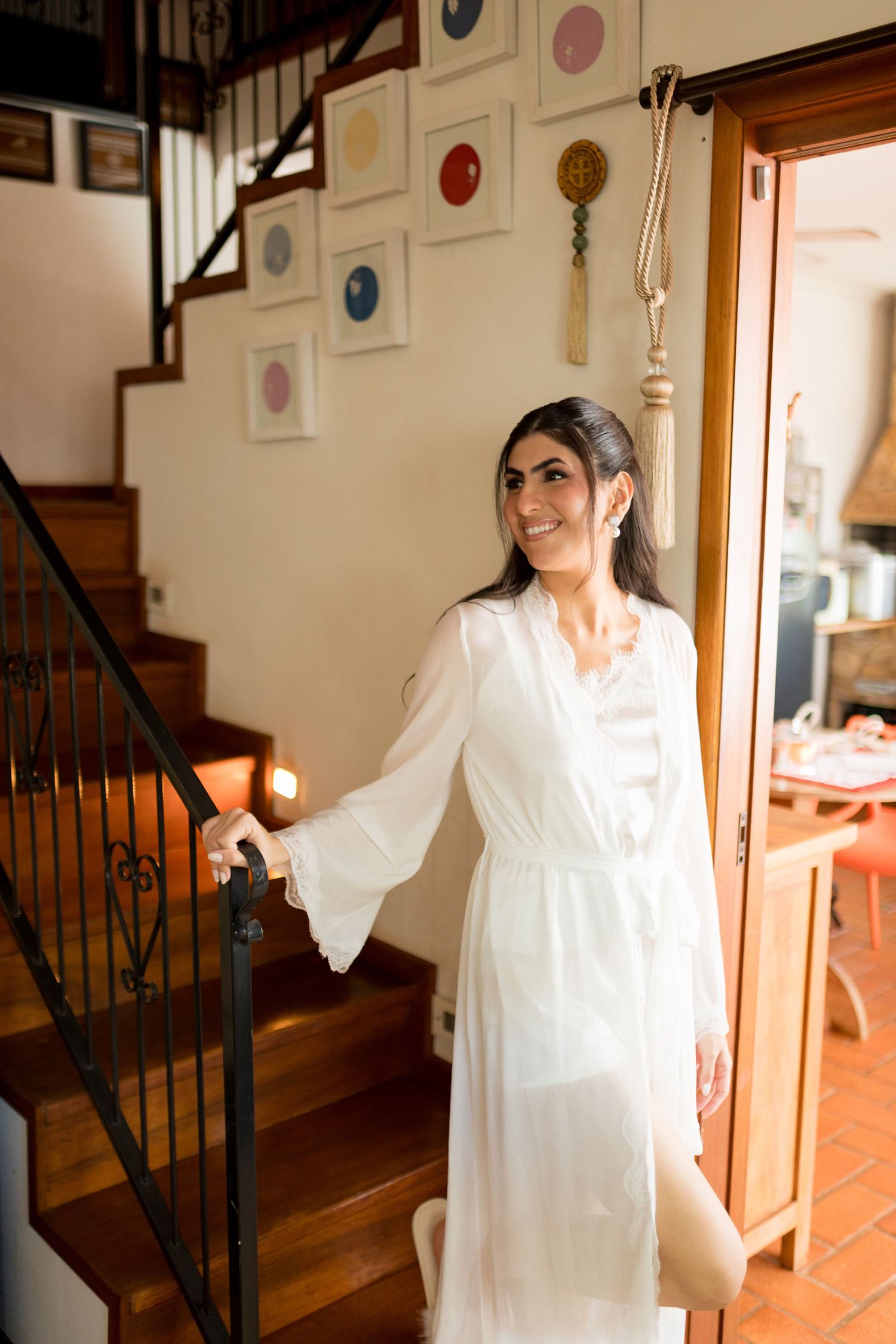 A smiling woman in a white dress stands by a staircase railing in an elegant home interior.
