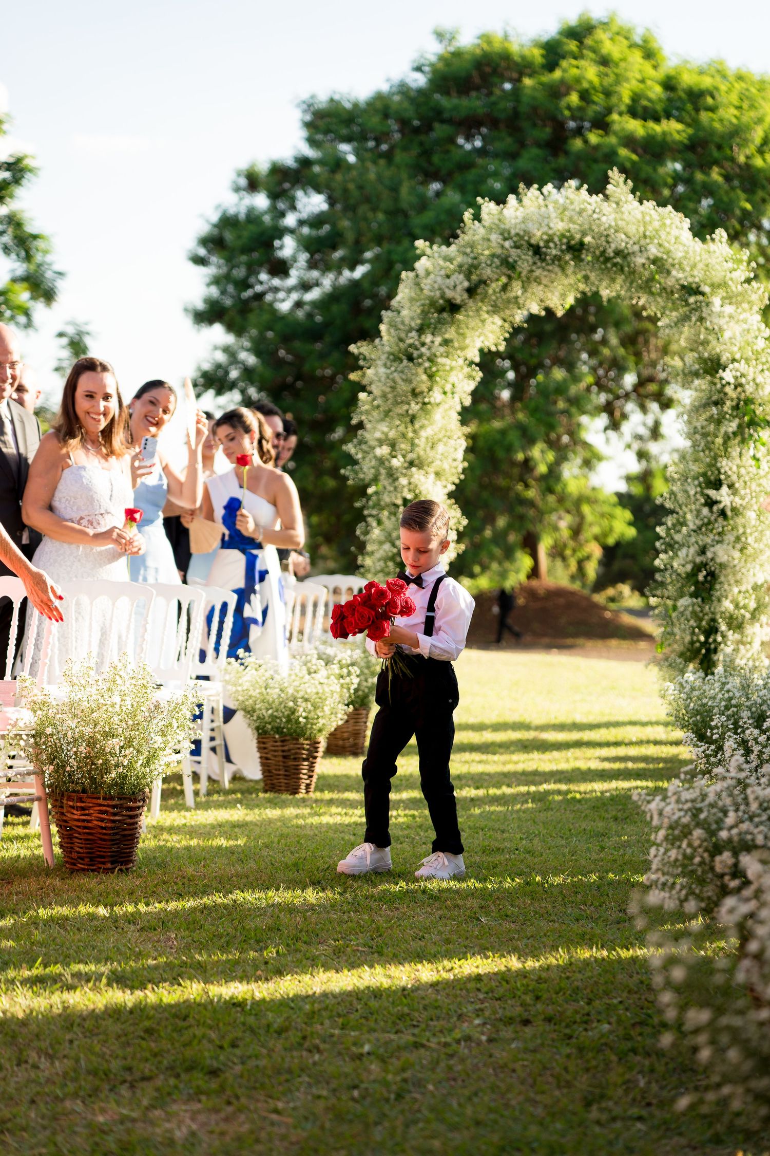Young ring bearer in black pants and red vest carries red roses down floral arch aisle at outdoor wedding ceremony.