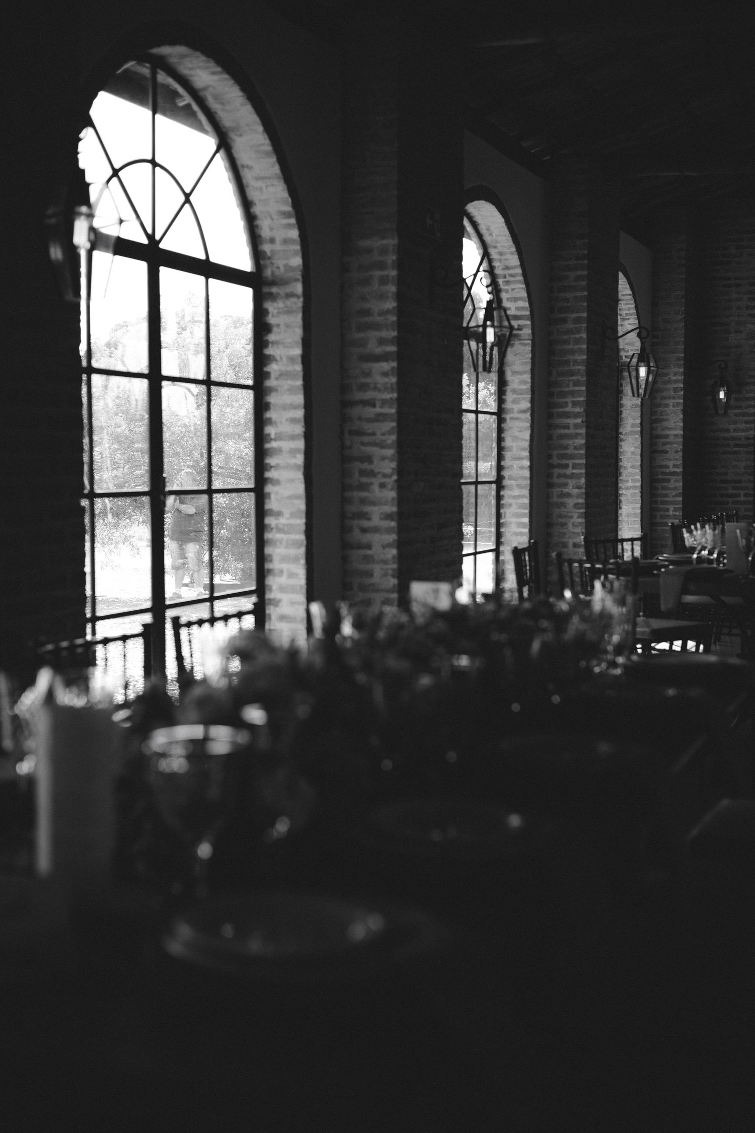 Black and white photo of elegant brick venue with arched windows, set tables in foreground, moody atmospheric lighting.
