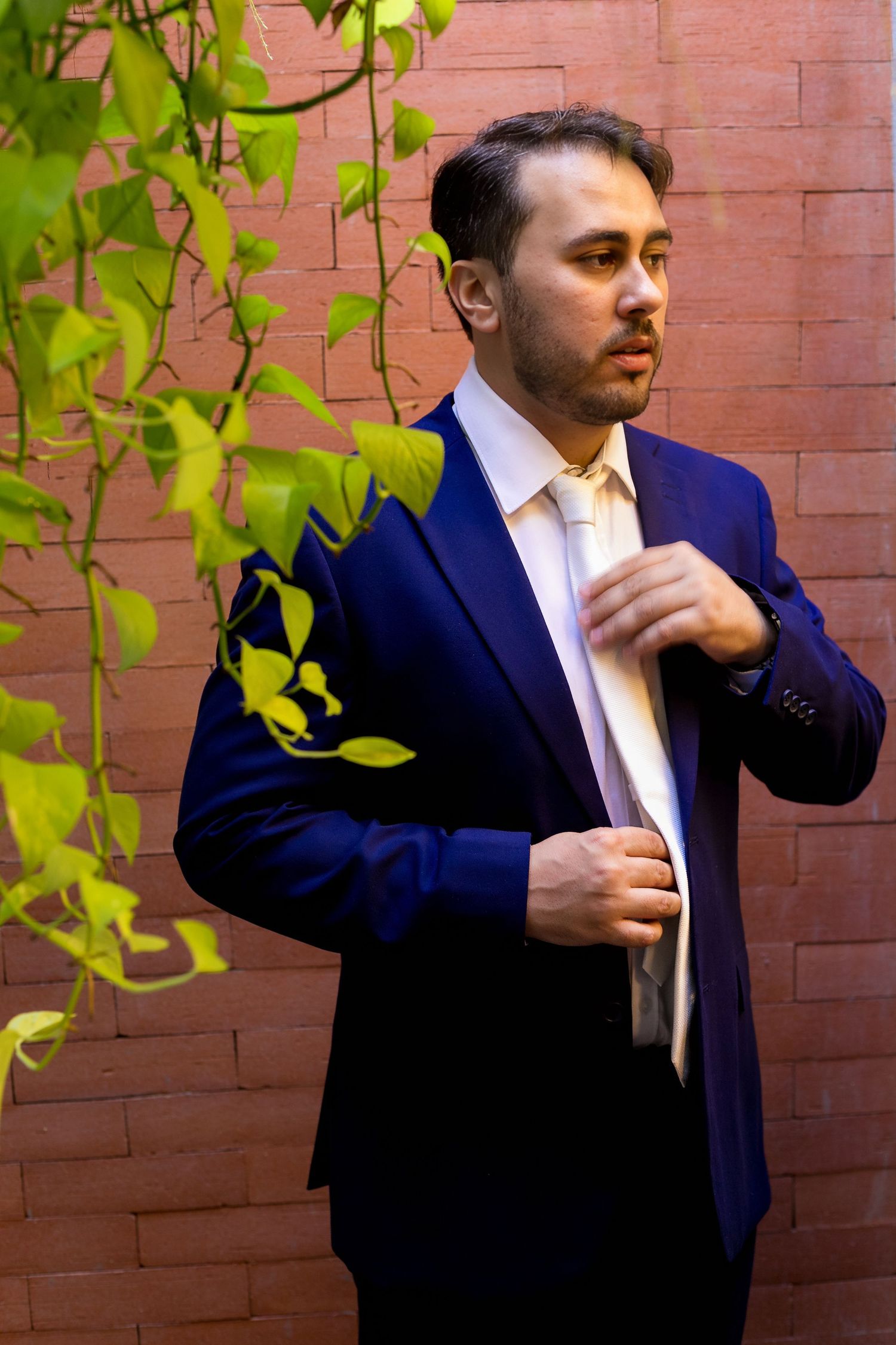 A well-dressed man in a navy blue suit adjusts his cream tie against a red brick wall with green ivy leaves.