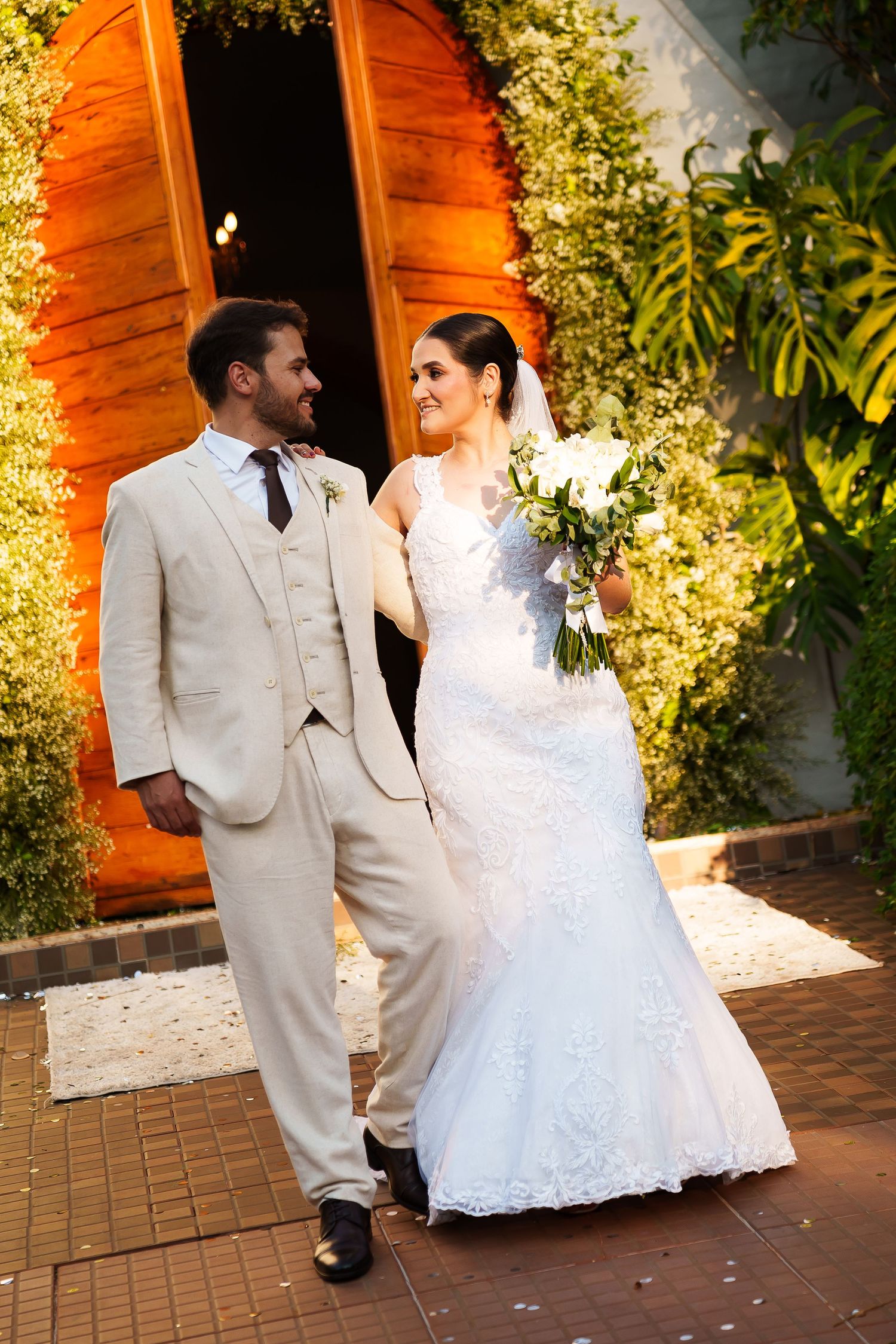 Bride in white lace gown holding bouquet walks with groom in beige suit outside decorated wedding venue.