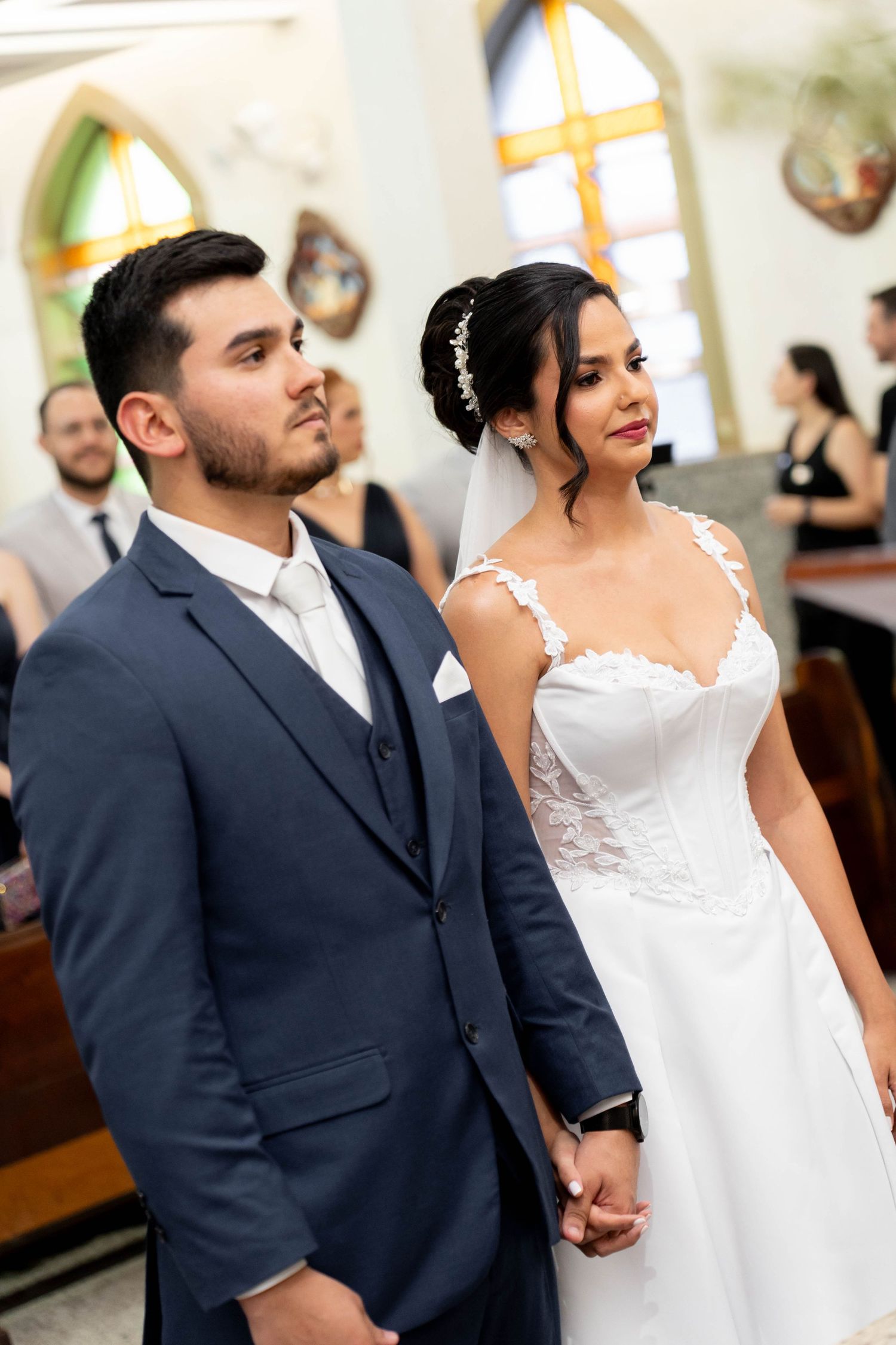 Bride and groom holding hands at church altar, she in lace gown with veil, he in navy blue suit.