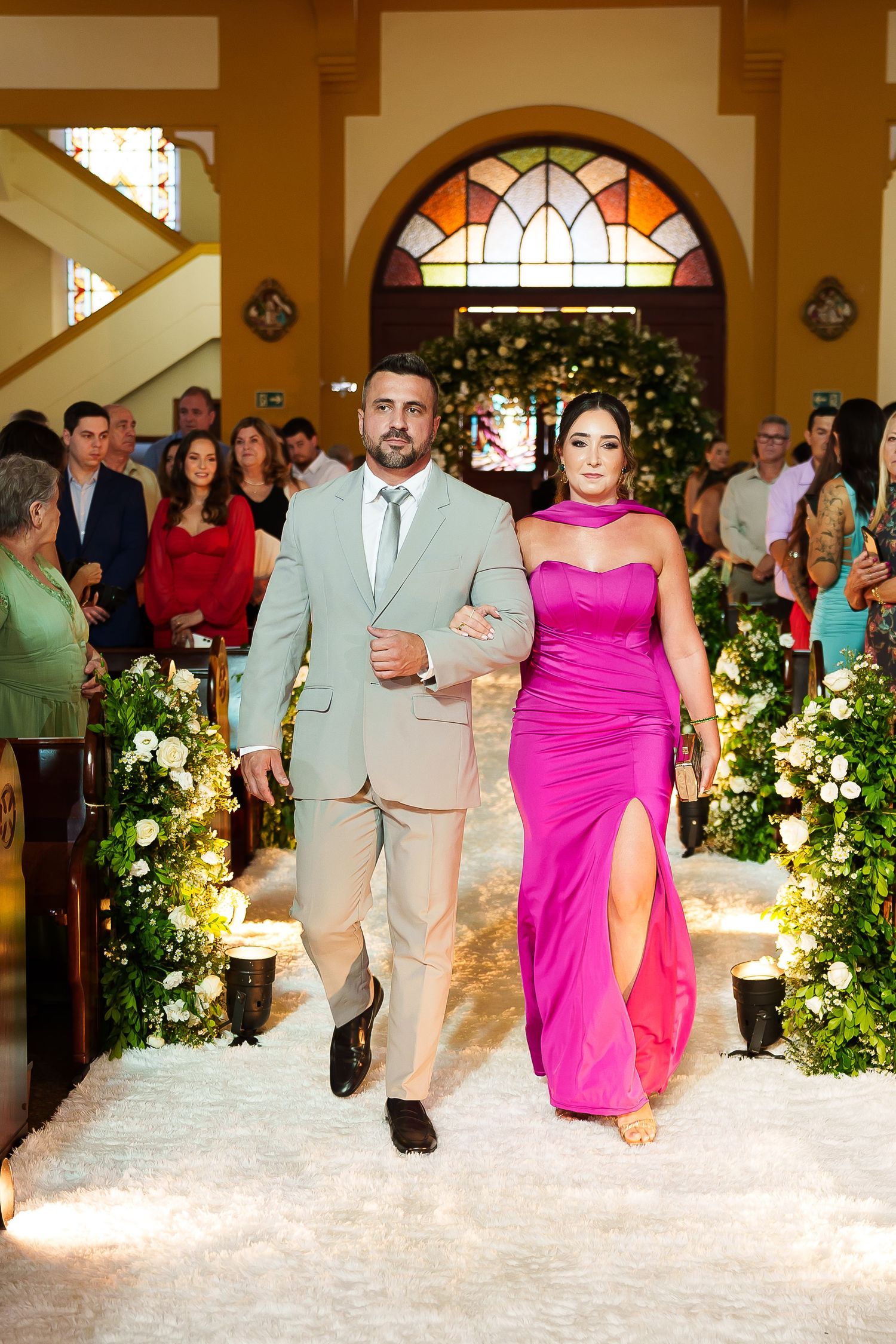 A couple walks down a church aisle, man in tan suit and woman in pink gown, during a wedding ceremony.