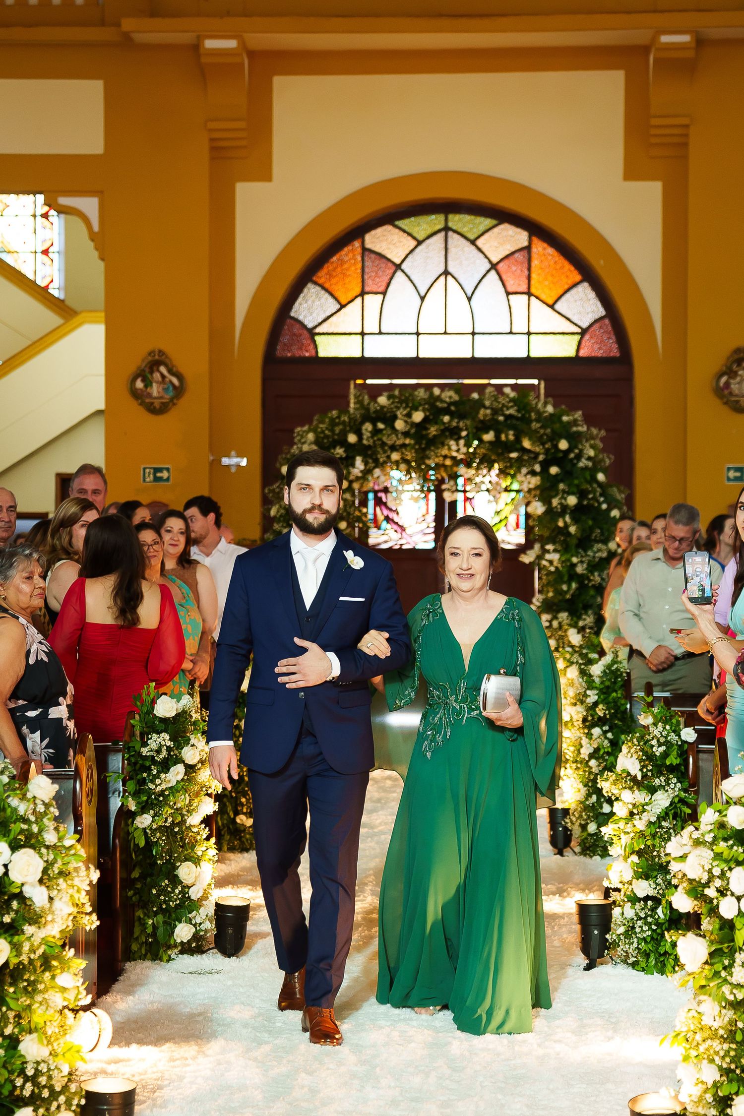 Groom in navy suit escorts woman in emerald green gown down floral-lined wedding aisle in elegant church.