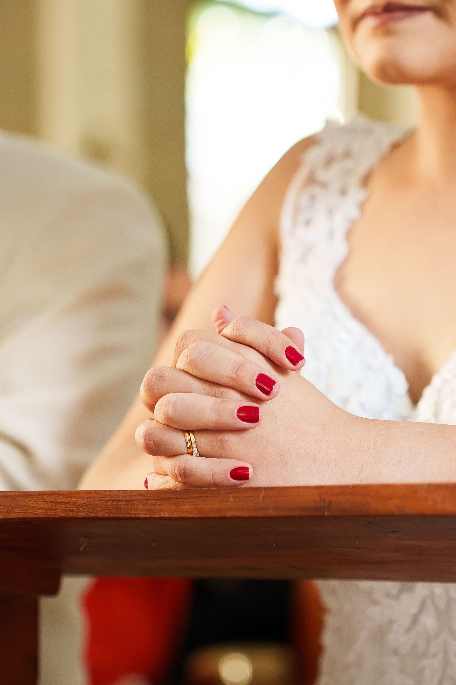 Bride in white lace dress with red nails and gold wedding ring clasping hands on wooden surface.