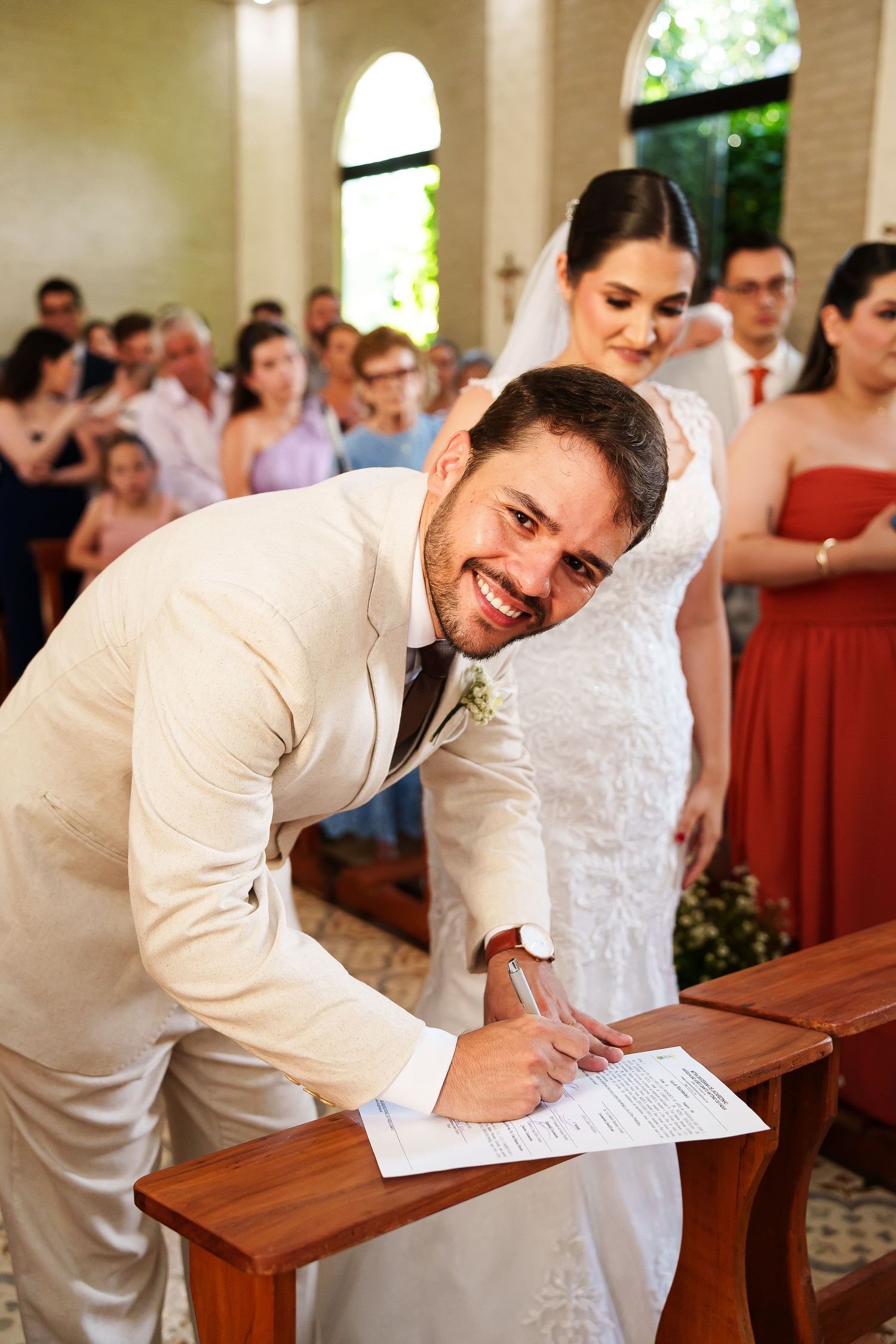 Smiling groom signs wedding certificate at altar while bride in white gown stands behind him in church.