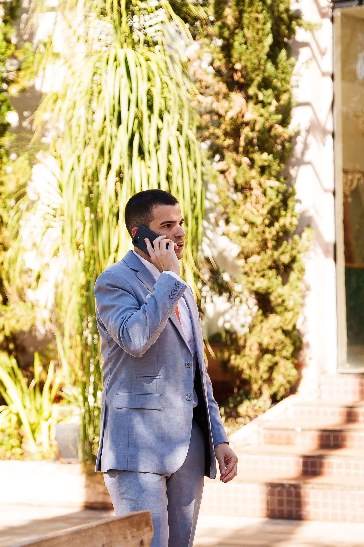Smiling man in light blue suit talking on phone outdoors near tropical plants and cacti.