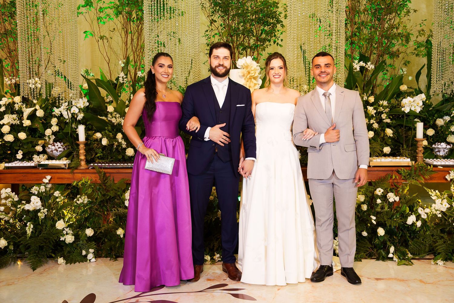 Four elegantly dressed guests pose together at a wedding reception with floral decorations in the background.