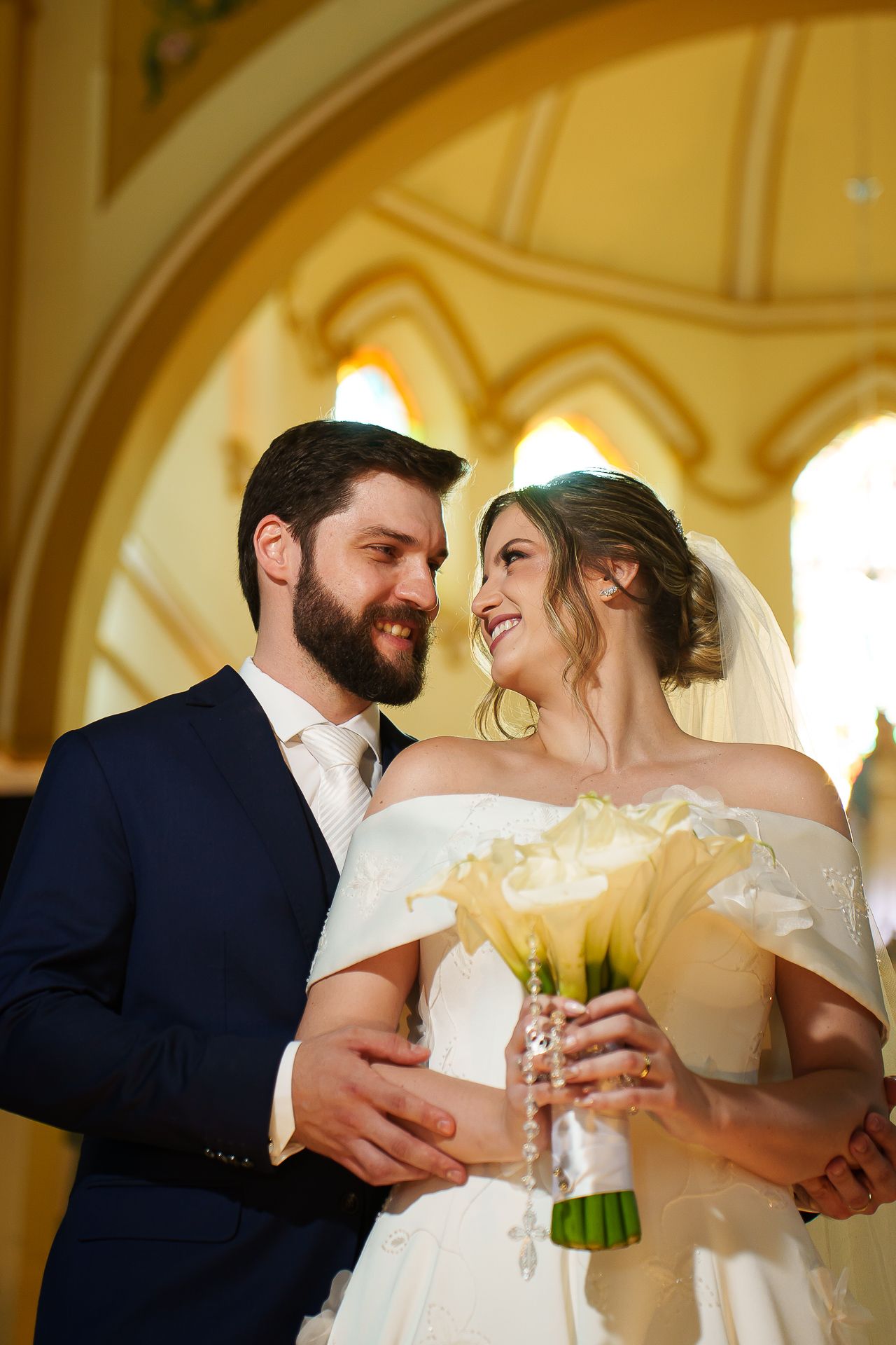 Smiling bride and groom embrace in golden church, bride holds white calla lily bouquet wearing off-shoulder gown.