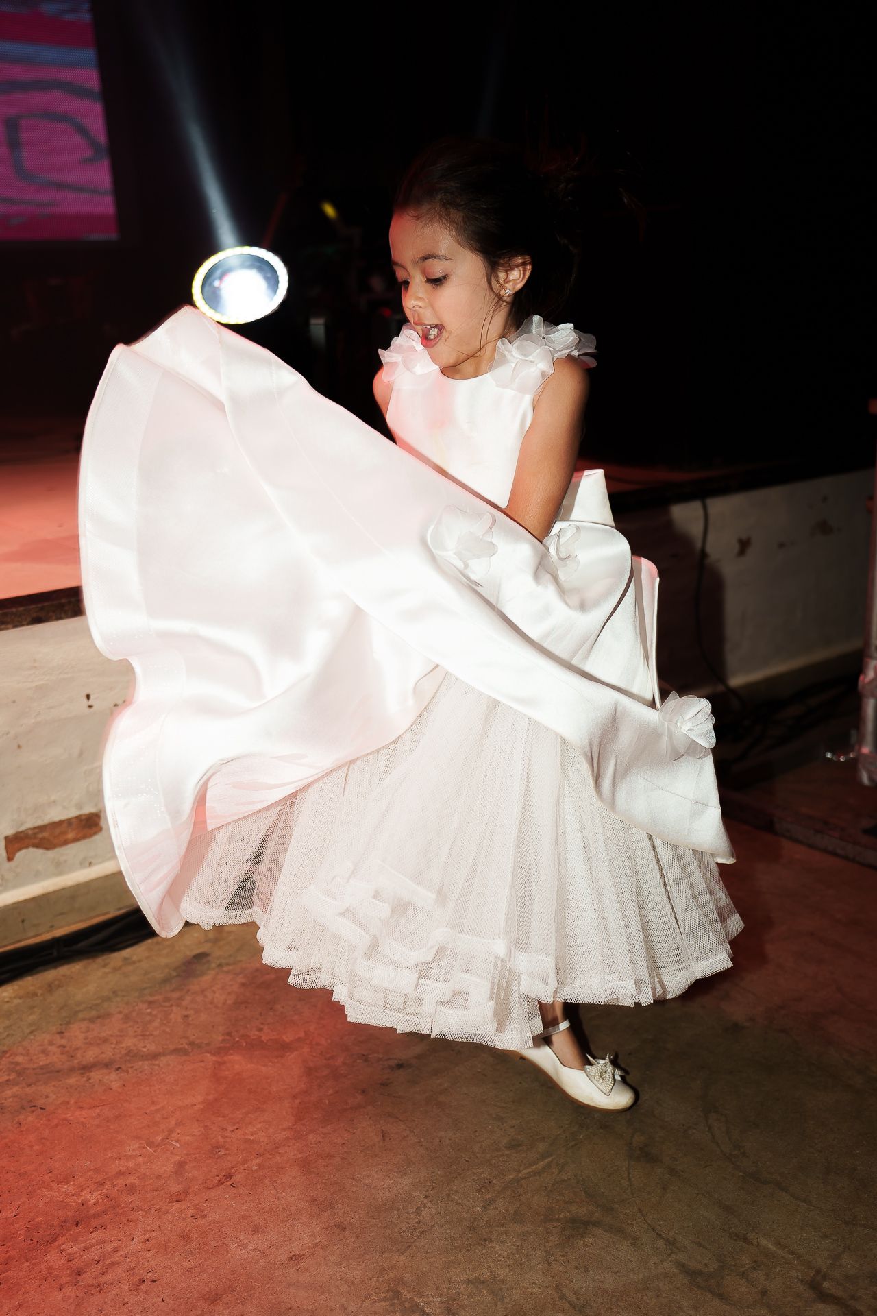 Young girl in white dress and gloves twirling on stage under spotlight at formal event.