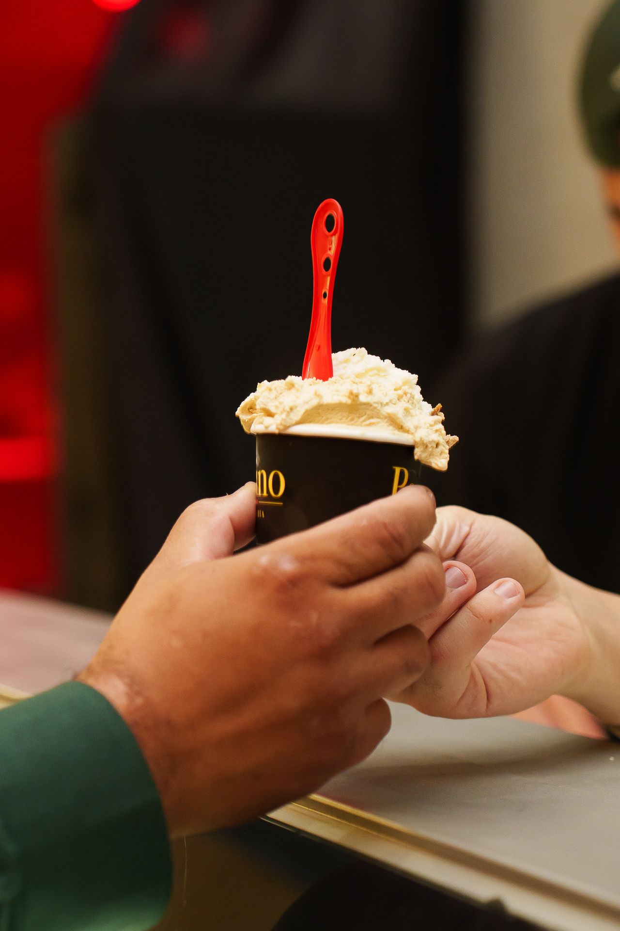 Two hands exchanging a black cup of creamy gelato topped with a red spoon at an ice cream shop.