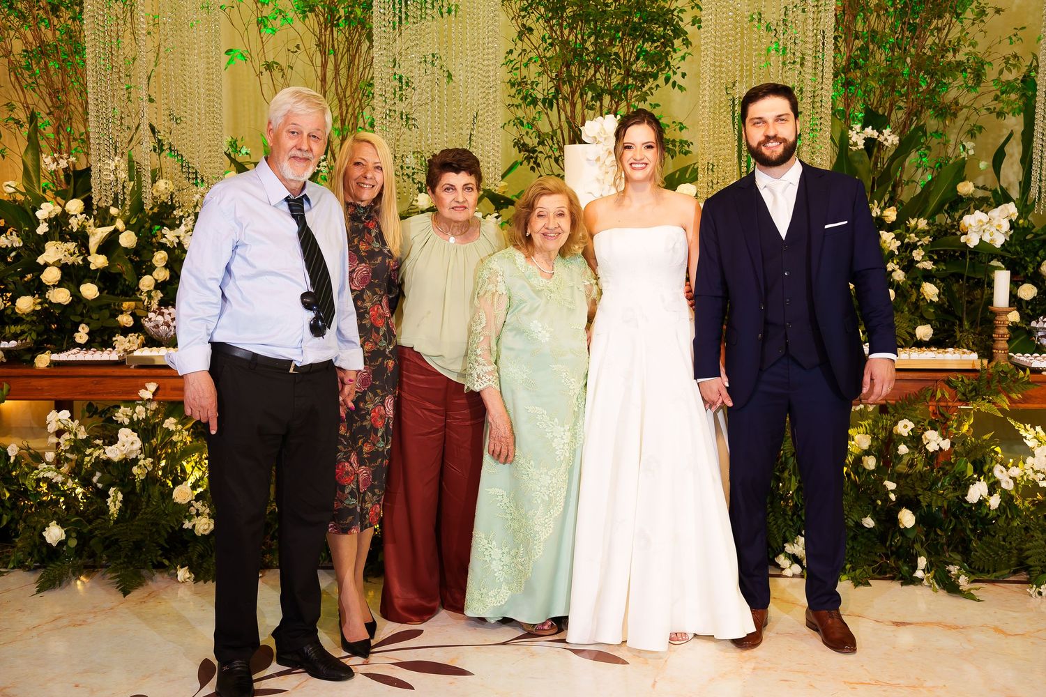 Wedding group photo with bride in white gown, groom in navy suit, and four family members posing before floral backdrop.