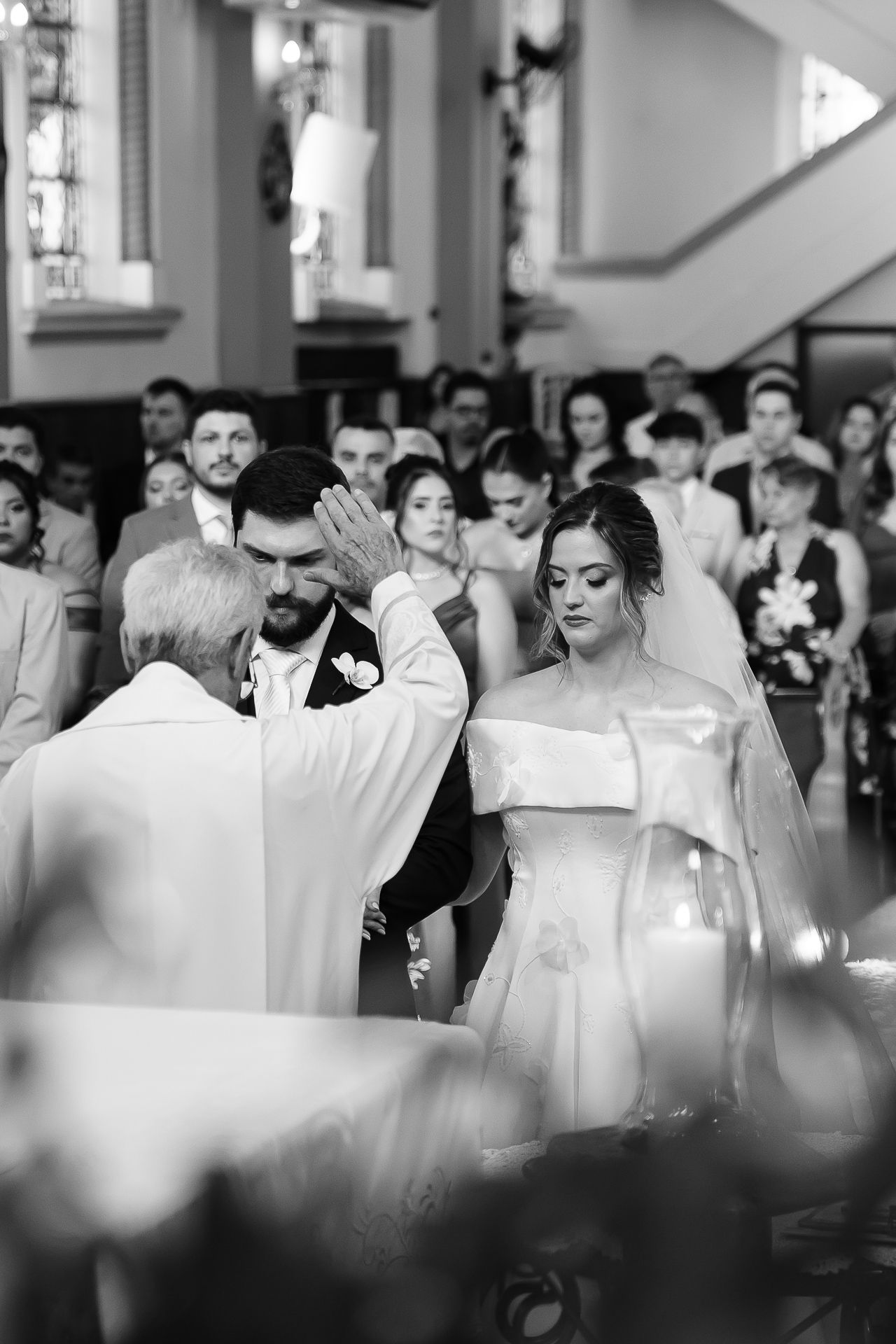 Black and white photo of a priest blessing a bride and groom during a church wedding ceremony with guests seated behind.