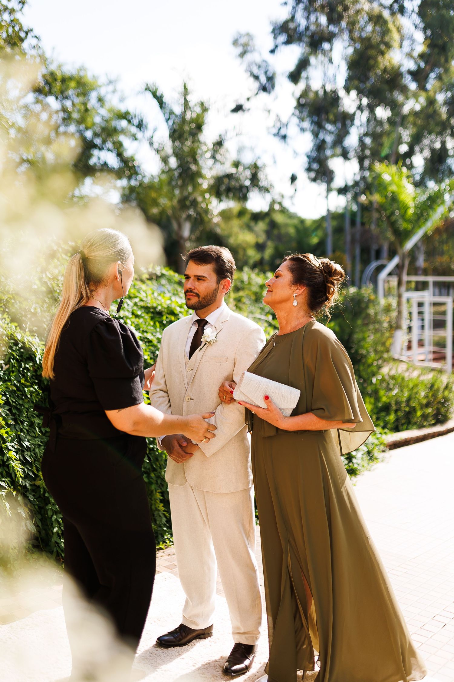 Elegant wedding guests in formal attire conversing outdoors, man in cream suit with women in black and olive green dresses.