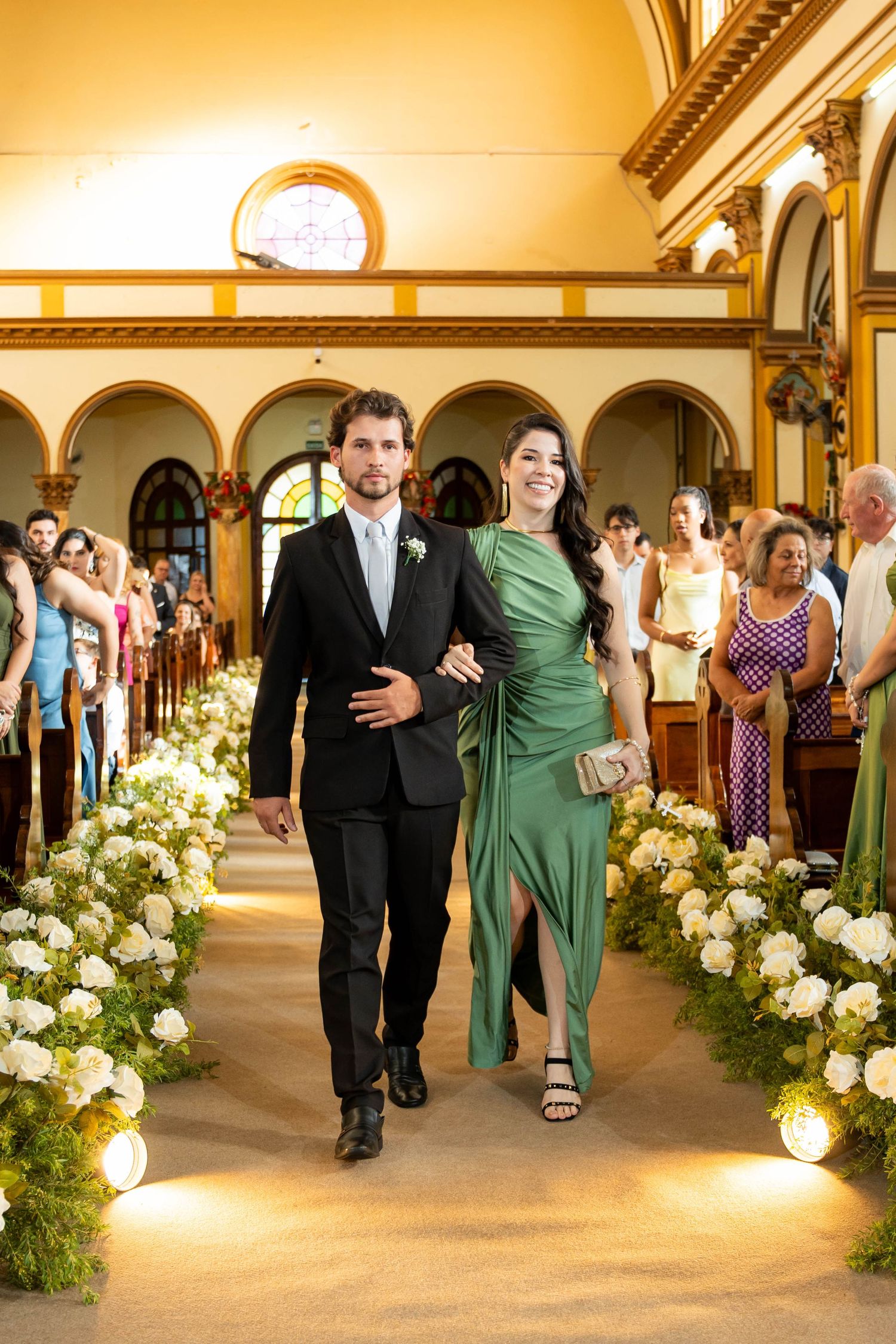 Couple walks down church aisle, man in black tuxedo, woman in green gown, flanked by white floral arrangements.