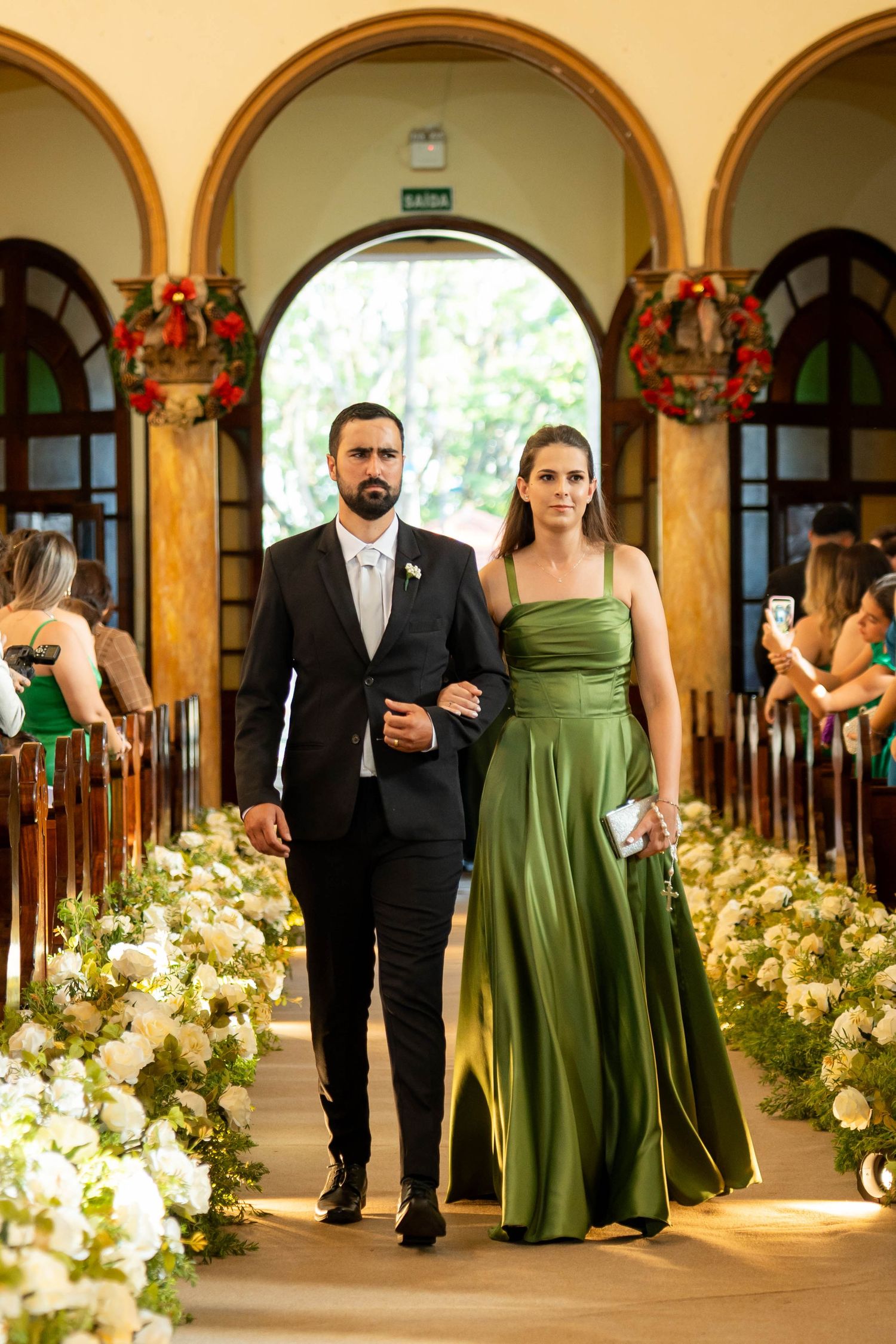 A couple walks down a flower-lined church aisle, man in black suit, woman in elegant green satin gown.