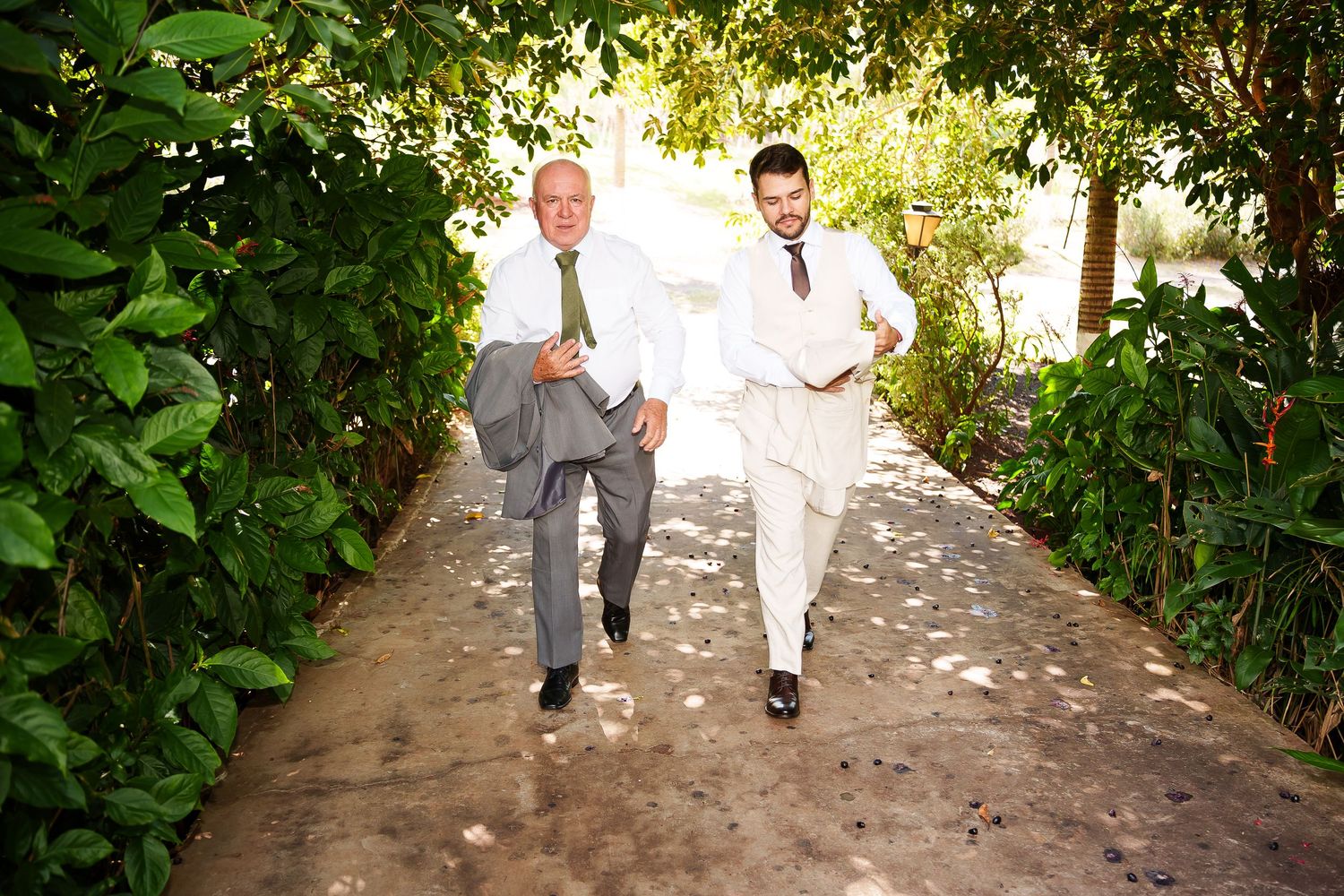 Two men in suits run joyfully down a garden path lined with lush green trees at a wedding celebration.