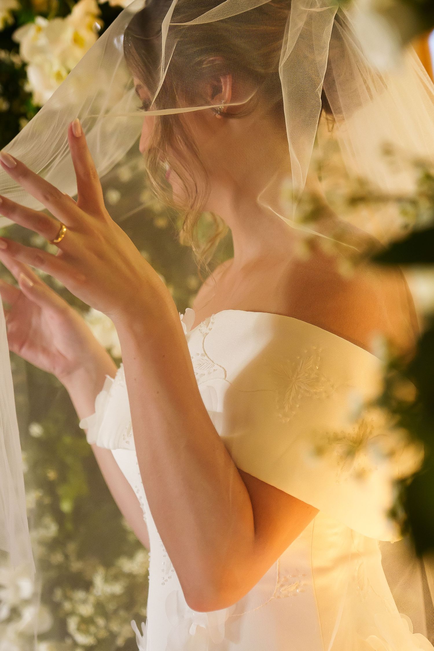 Bride in white strapless gown and veil, adorned with a gold ring, poses gracefully among lush floral arrangements.