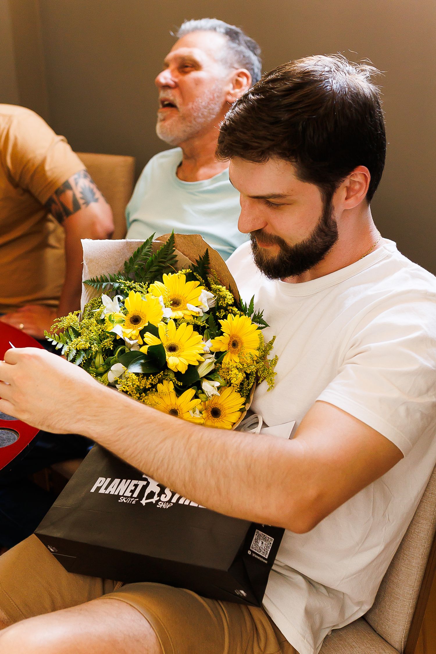 Young bearded man holding yellow flower bouquet with Planet Stars gift bag, seated beside older tattooed man.