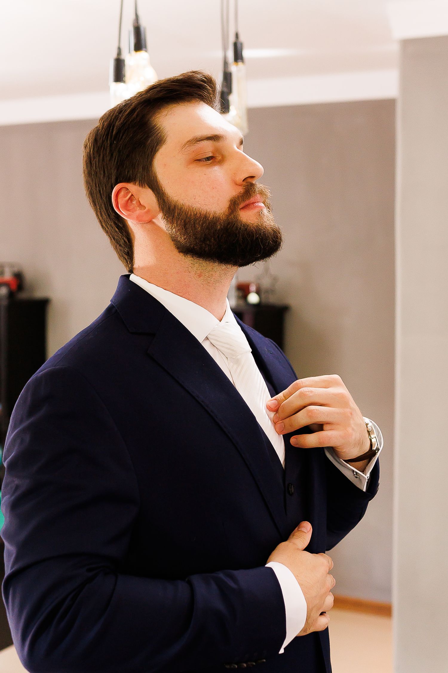 Bearded man in navy blue suit adjusting his tie, standing confidently indoors with eyes closed.