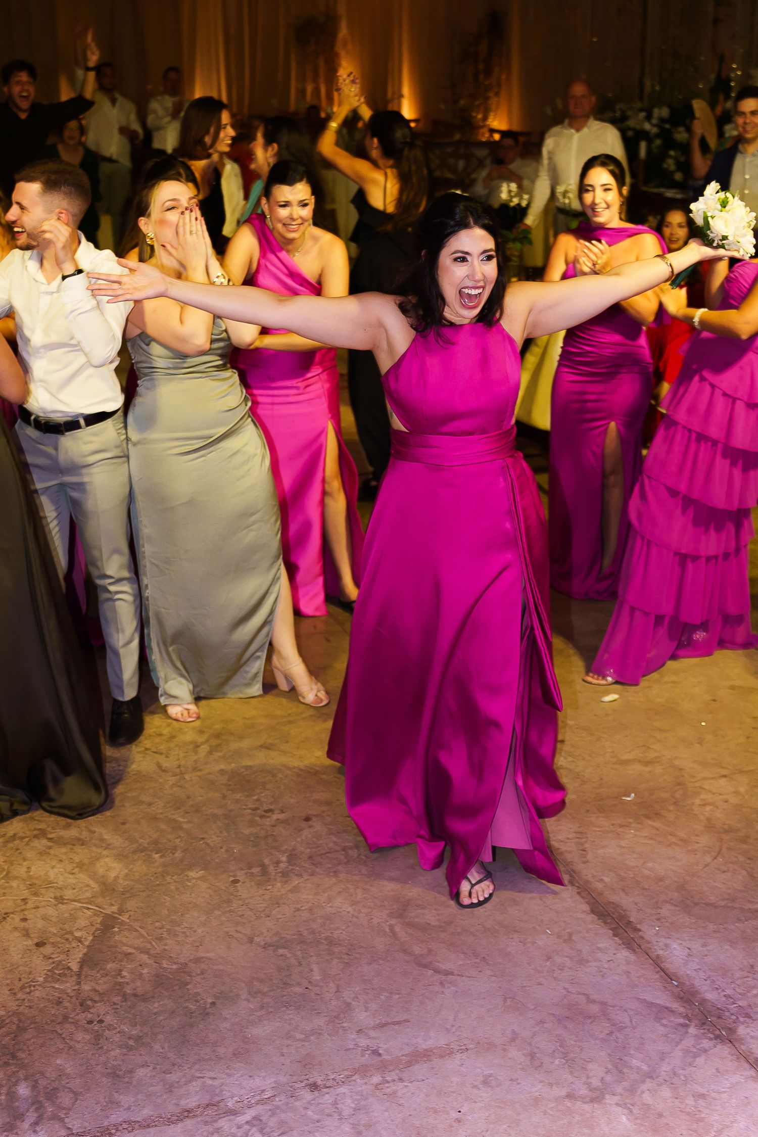 Bridesmaids in fuchsia gowns dance joyfully at a wedding reception, with one woman leading the group with arms outstretched.