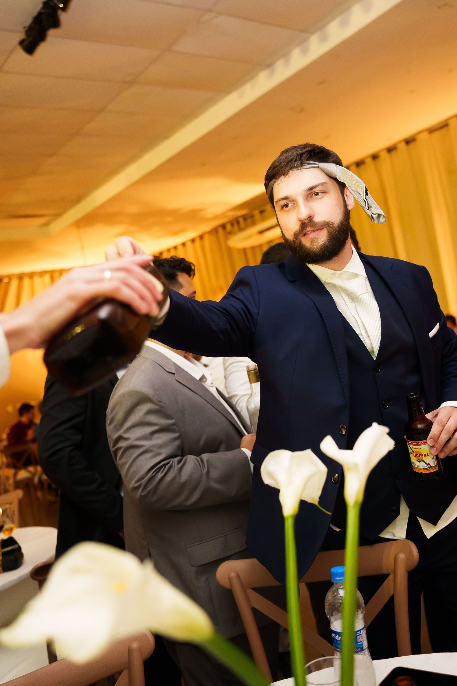 Bearded groom in navy suit reaching for champagne bottle at elegant wedding reception with white calla lilies.
