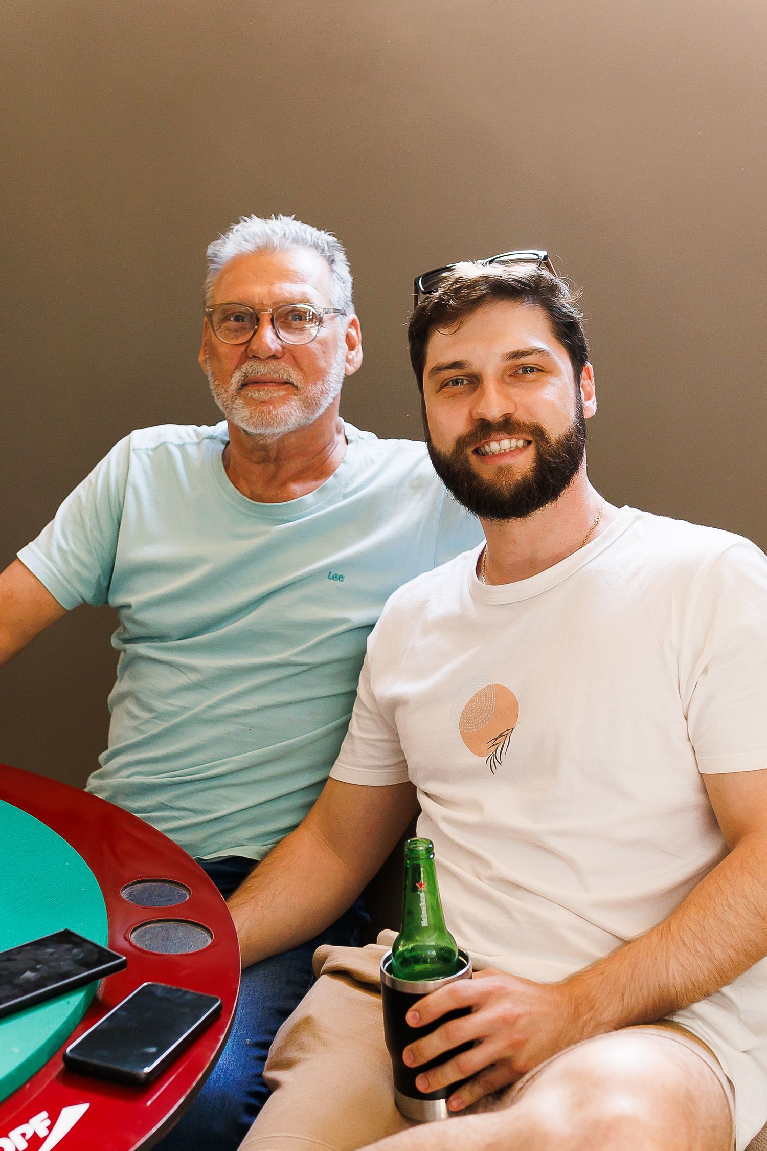Two men smiling at a table, one older with gray hair and glasses, one younger with a beard holding a green beer bottle.