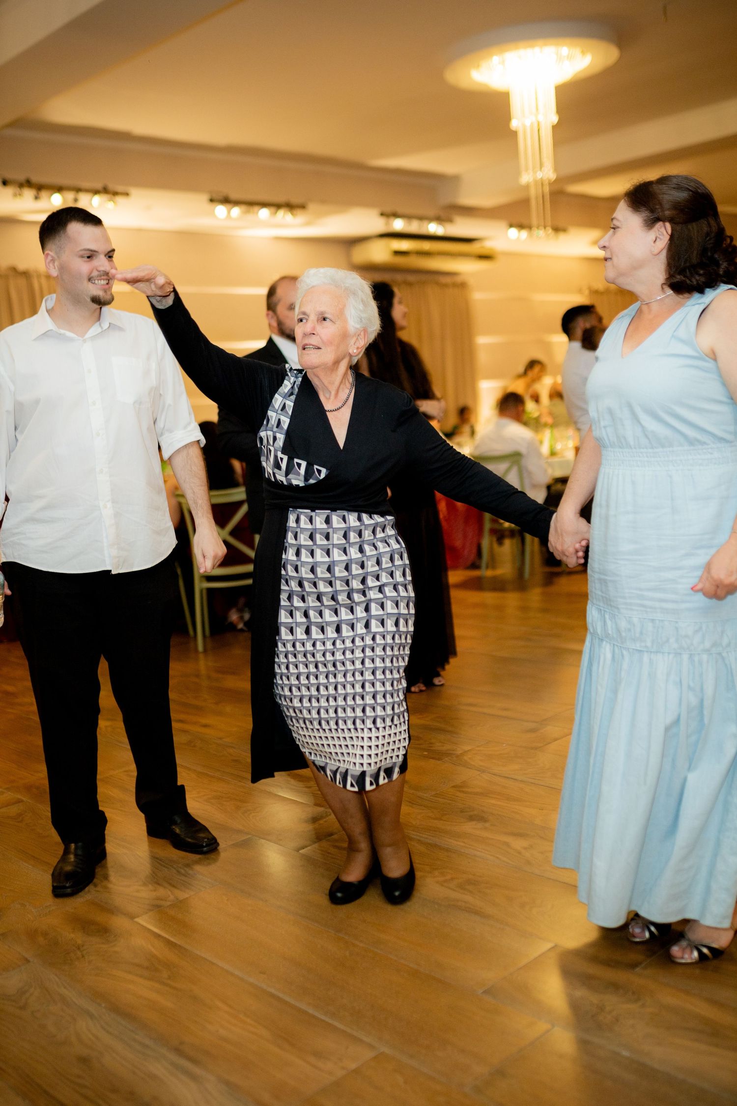 Elderly woman dancing joyfully with younger man and woman at a formal event on a wooden dance floor.