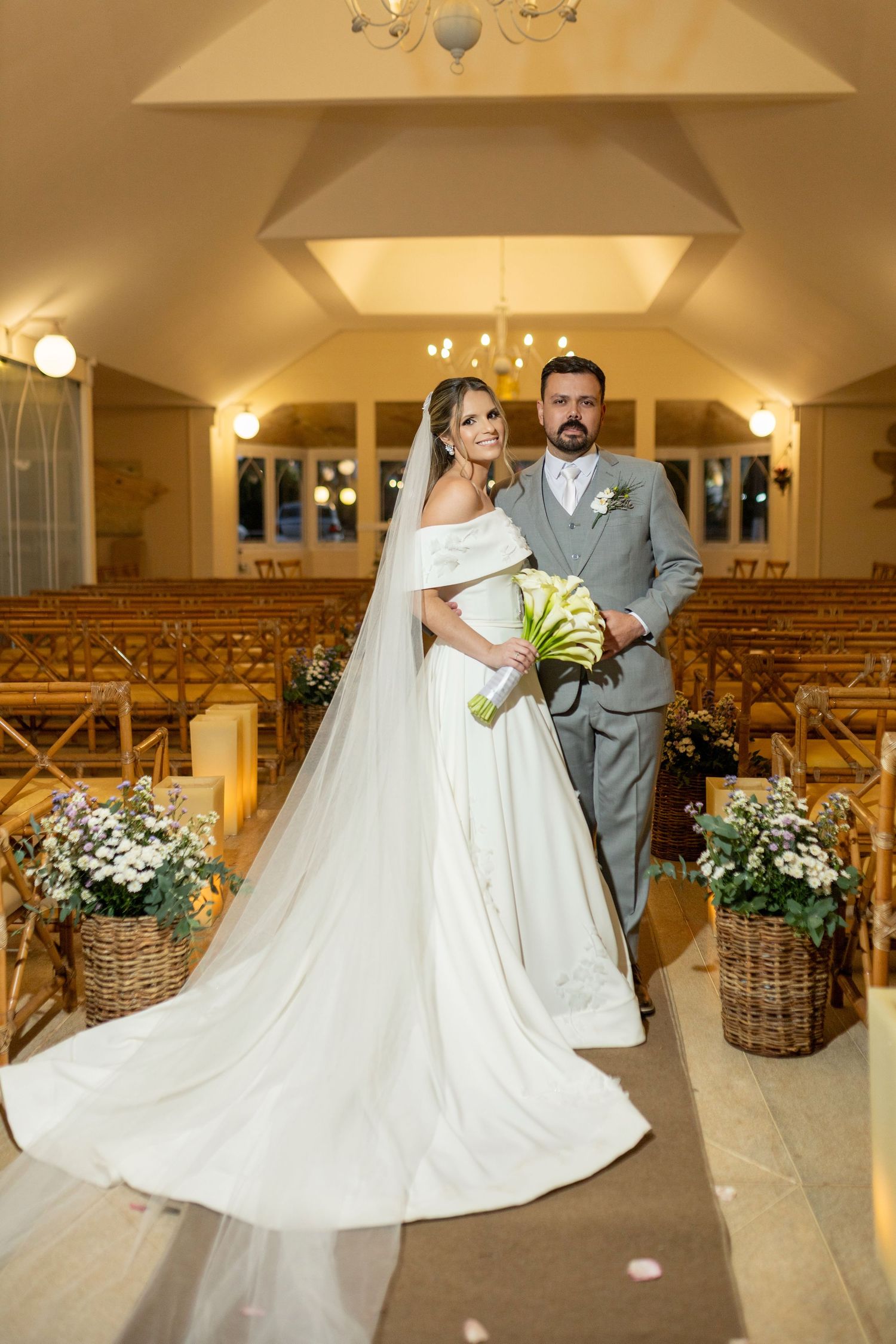Bride and groom pose elegantly in a warmly lit chapel aisle, bride in white gown with veil, groom in gray suit.
