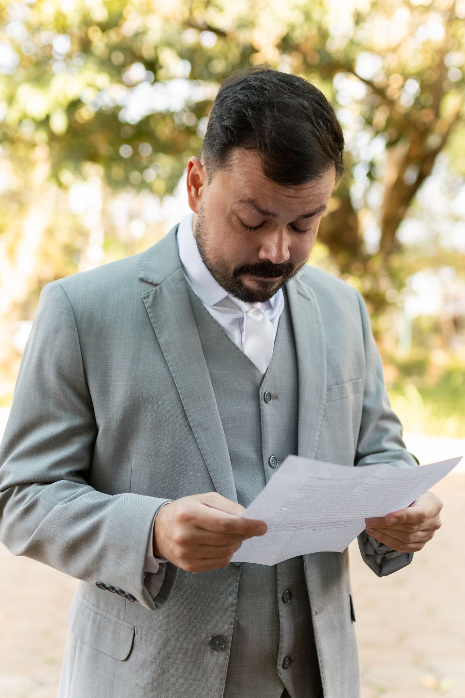 A bearded man in a gray suit reads a document outdoors with blurred green trees in the background.