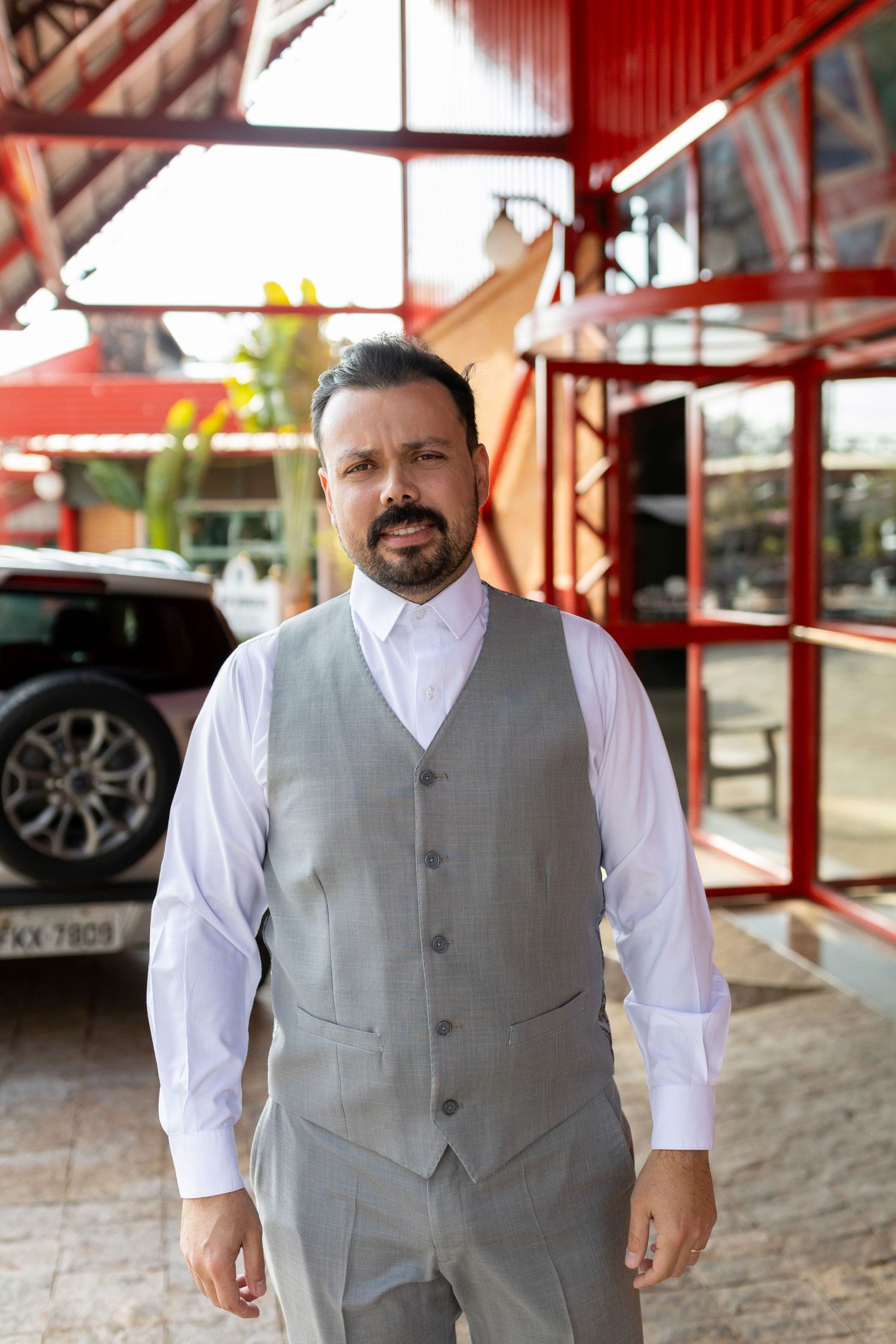 A bearded man in a gray vest and white dress shirt stands confidently outside a building with red metal framing.