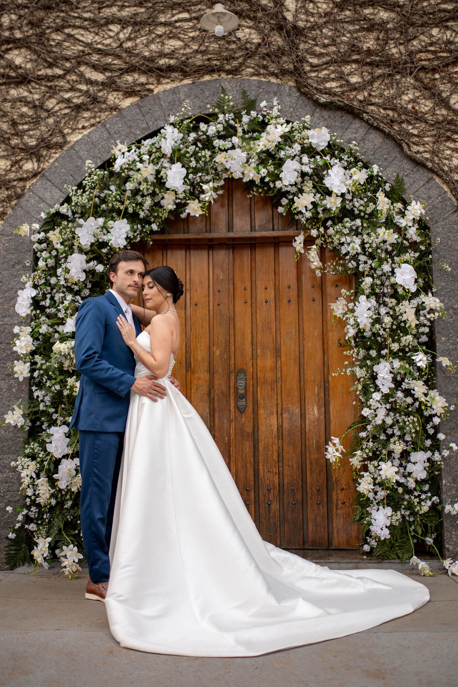 Bride and groom embrace under a floral arch of white flowers and greenery before a wooden door on their wedding day.