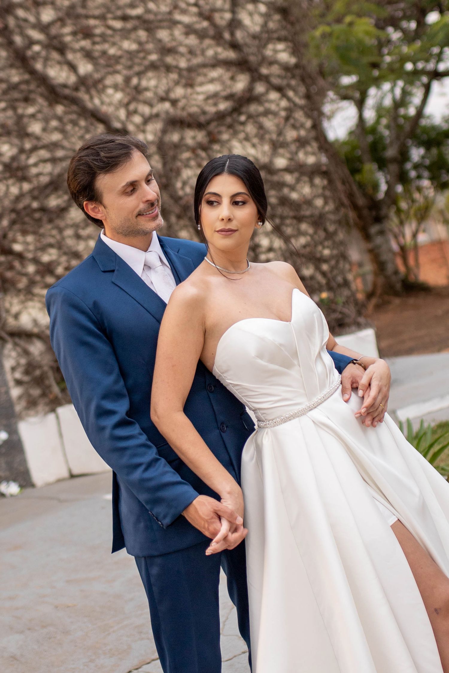 Bride in strapless white gown and groom in blue suit pose together outdoors in a romantic wedding portrait.