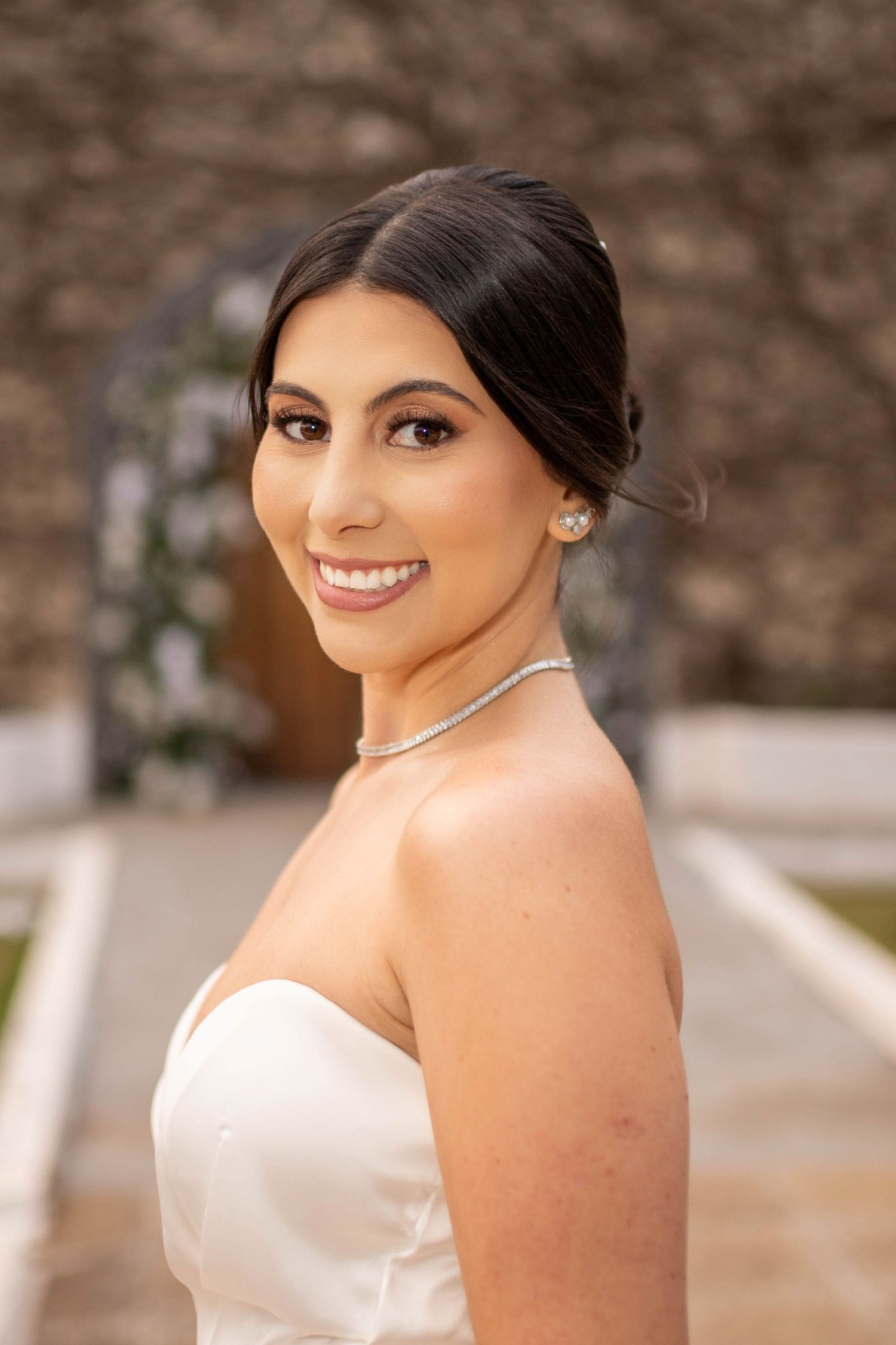 Smiling bride in strapless white gown with silver necklace and earrings, elegant updo hairstyle, outdoor wedding venue.