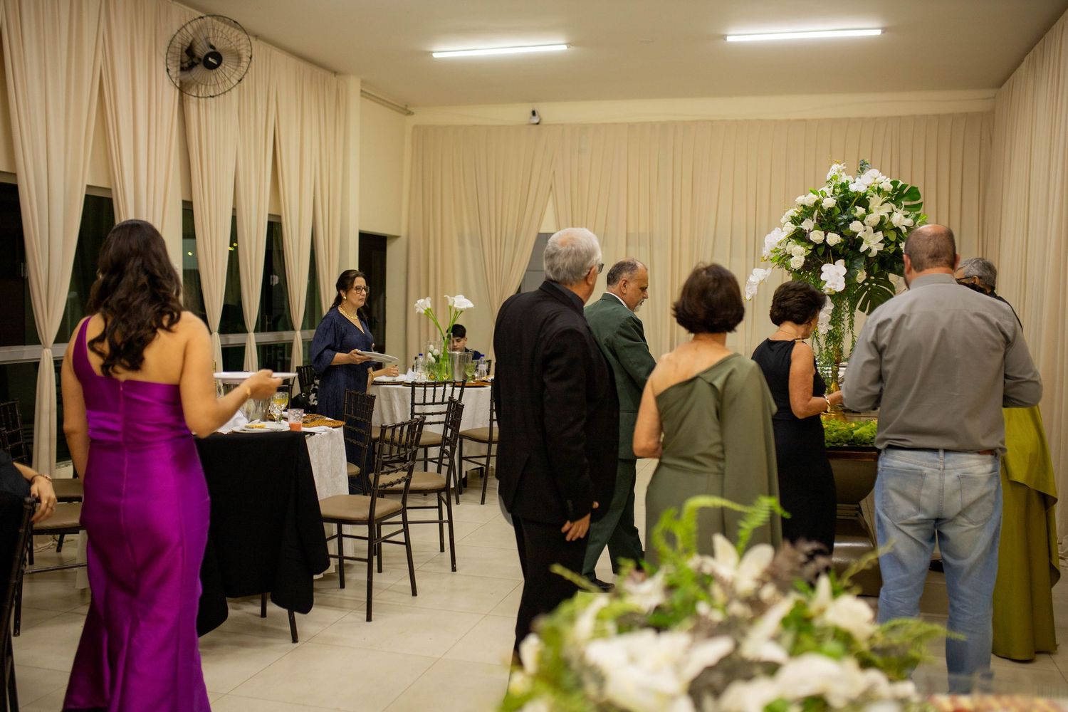 Elegant wedding reception with guests in formal attire, floral arrangements, and draped white curtains in a warmly lit venue.
